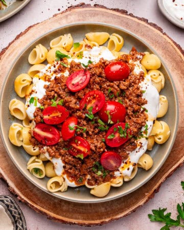 A plate of Turkish pasta, featuring shell pasta topped with ground meat sauce, yogurt, halved cherry tomatoes, and chopped herbs, served on a round wooden platter with parsley and a bowl of yogurt on the side.