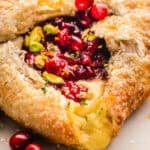 A close-up of a flaky puff pastry filled with melted brie cheese, bright red cranberries, and topped with chopped pistachios and sugar crystals. The crust is golden and slightly glossy.
