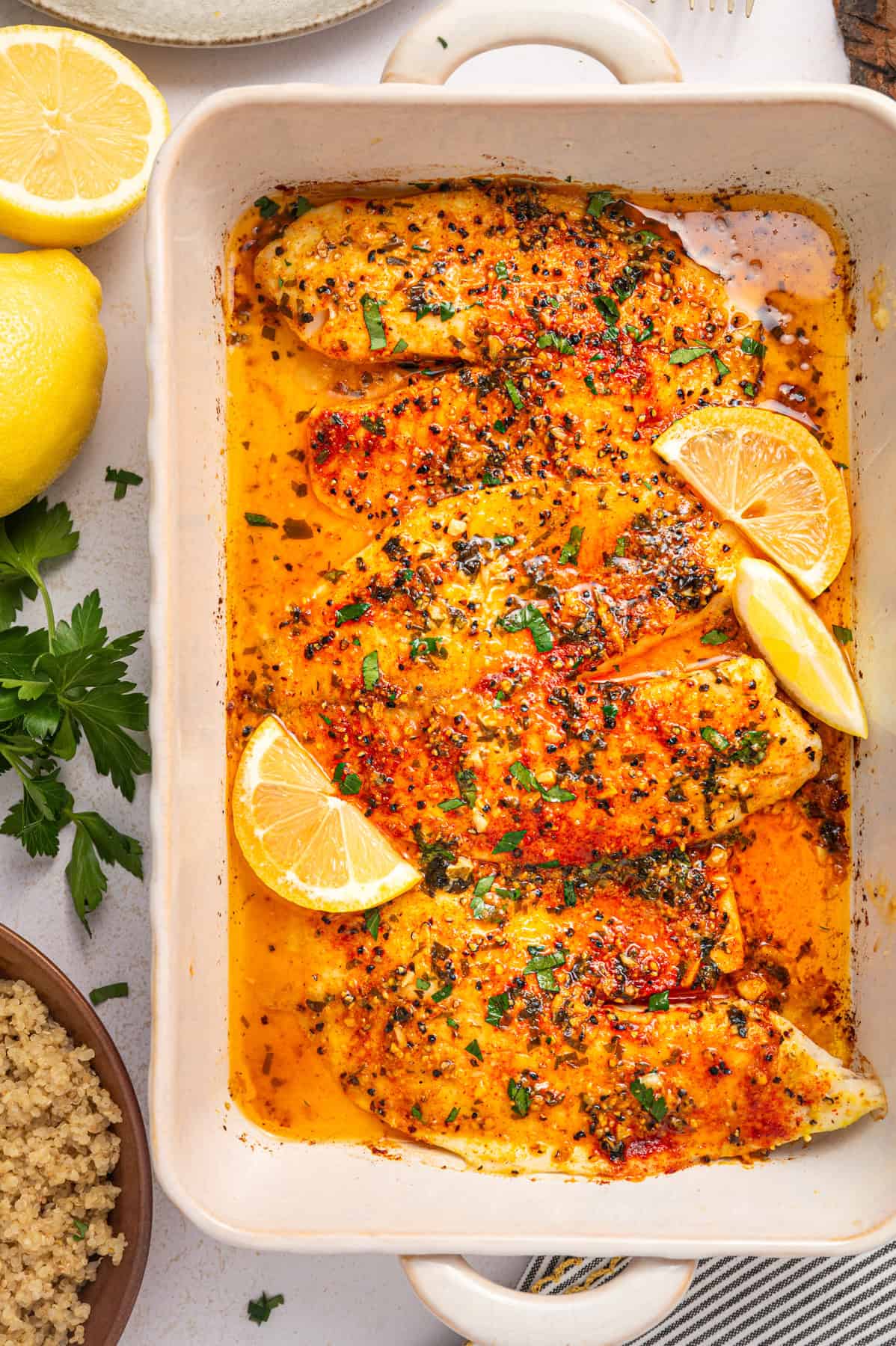 Lemon Pepper Baked Tilapia (with Lemon Butter Garlic Sauce) Baked tilapia fillets in a white dish, topped with herbs and spices in a golden, buttery sauce. Garnished with fresh parsley and lemon wedges. Lemons and parsley are nearby on the table.