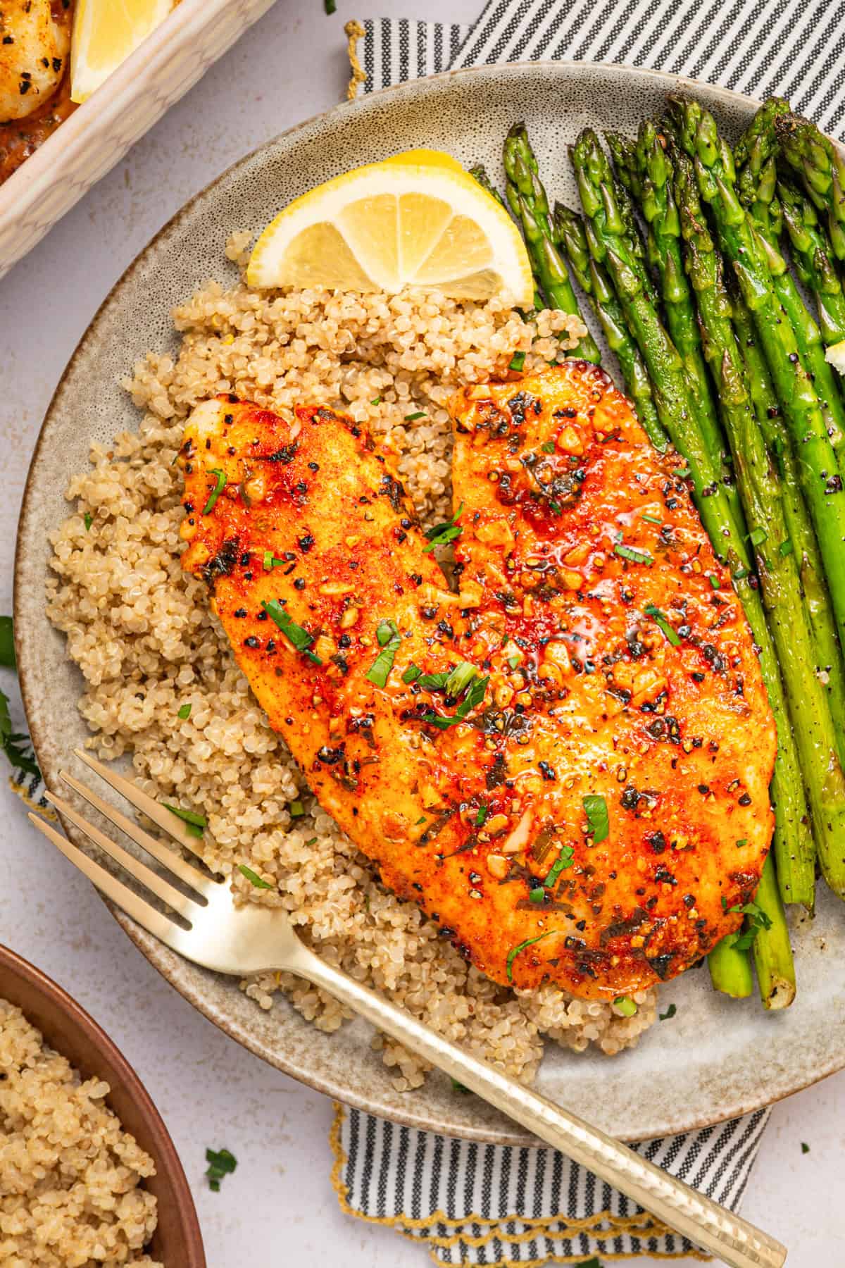 Lemon Pepper Baked Tilapia (with Lemon Butter Garlic Sauce) A plate with seasoned baked tilapia, cooked quinoa, and roasted asparagus, garnished with herbs and a lemon wedge. A fork rests on the plate, and a striped napkin is partially visible underneath.