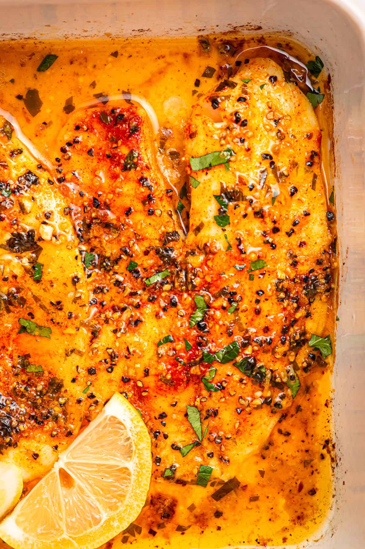 Lemon Pepper Baked Tilapia (with Lemon Butter Garlic Sauce) Close-up of baked seasoned tilapia fillets in a golden sauce, garnished with chopped herbs and cracked pepper, with a lemon wedge in the corner of the dish.