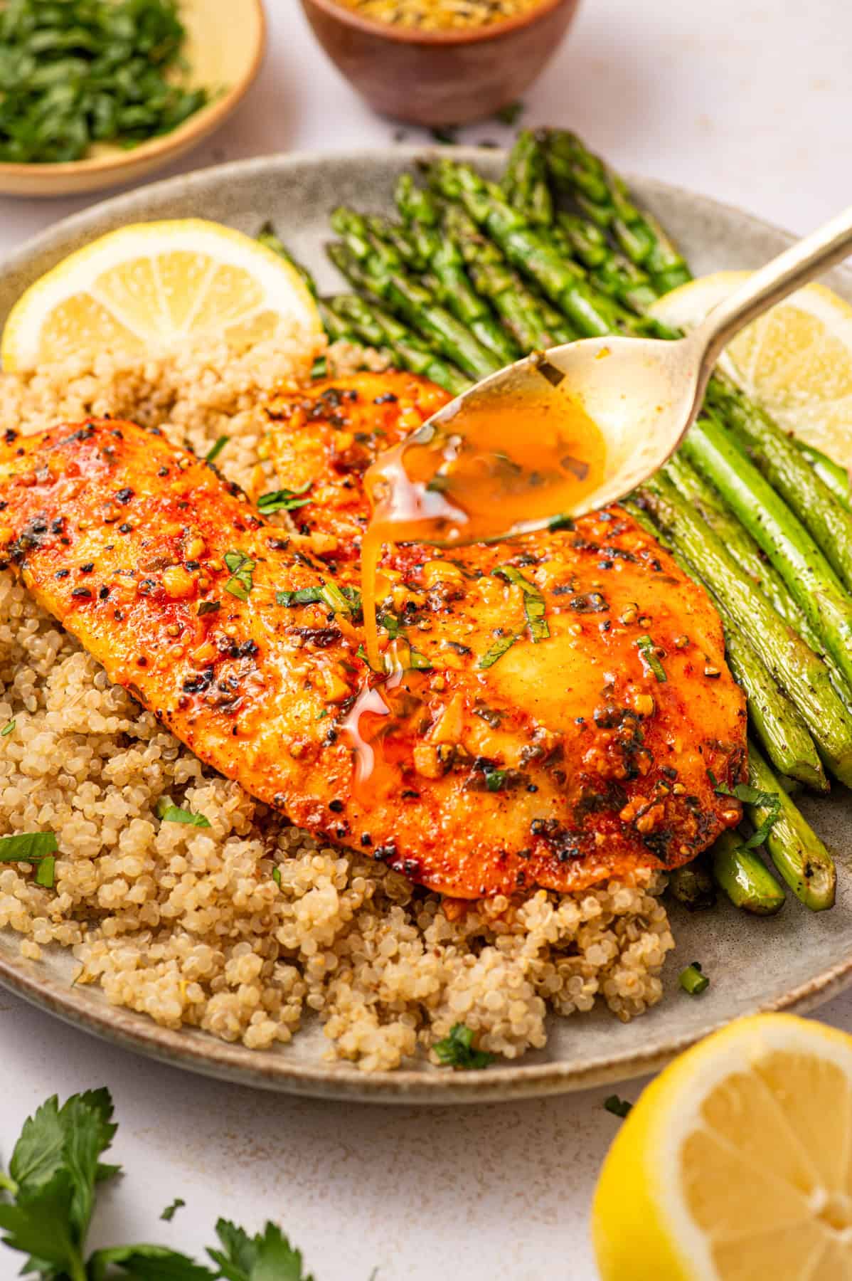 Lemon Pepper Baked Tilapia (with Lemon Butter Garlic Sauce) A plate with seasoned baked tilapia, quinoa, and roasted asparagus. A spoon pours sauce over the fish. Lemon slices and herbs garnish the plate, with extra greens and lemon in the background.