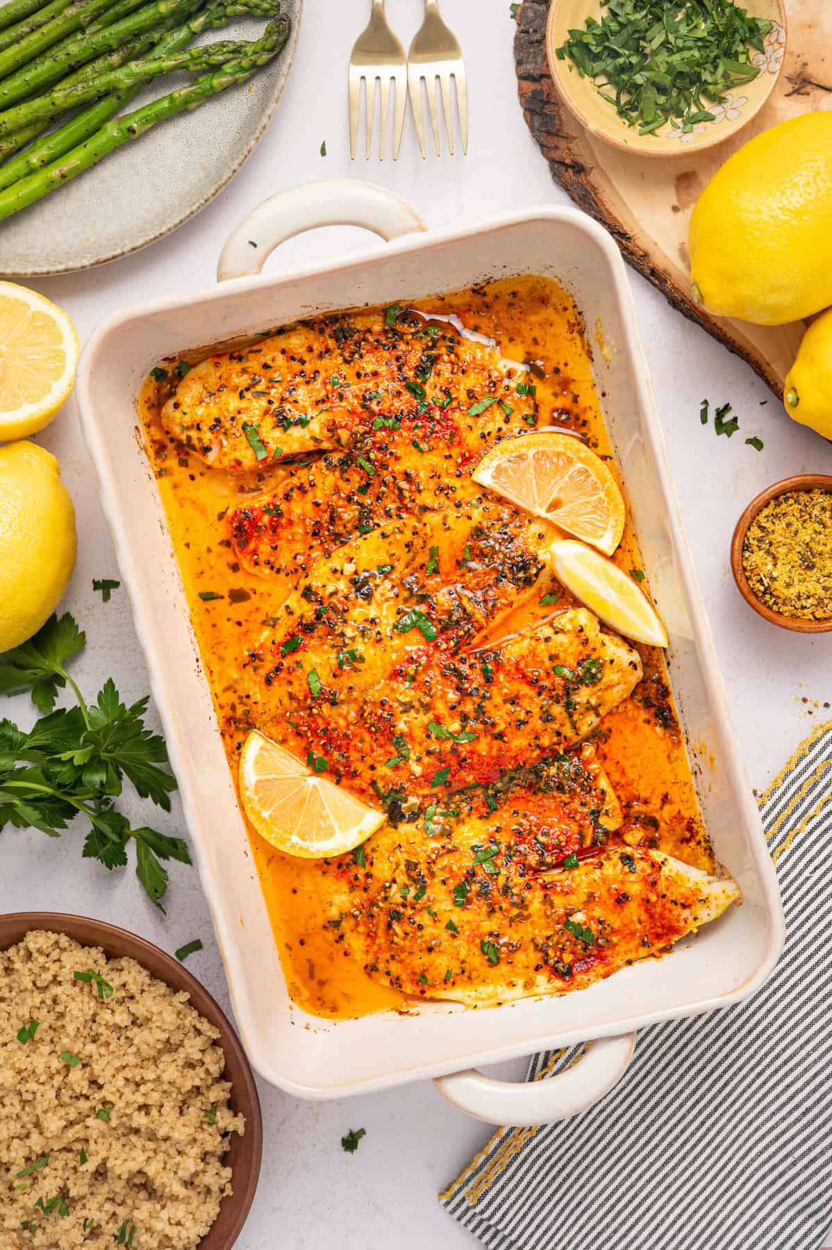 Lemon Pepper Baked Tilapia (with Lemon Butter Garlic Sauce) Baked tilapia fillets in a white dish with a golden, herbed sauce, garnished with lemon wedges and parsley. Surrounding the dish are lemons, asparagus, quinoa, herbs, and serving utensils.