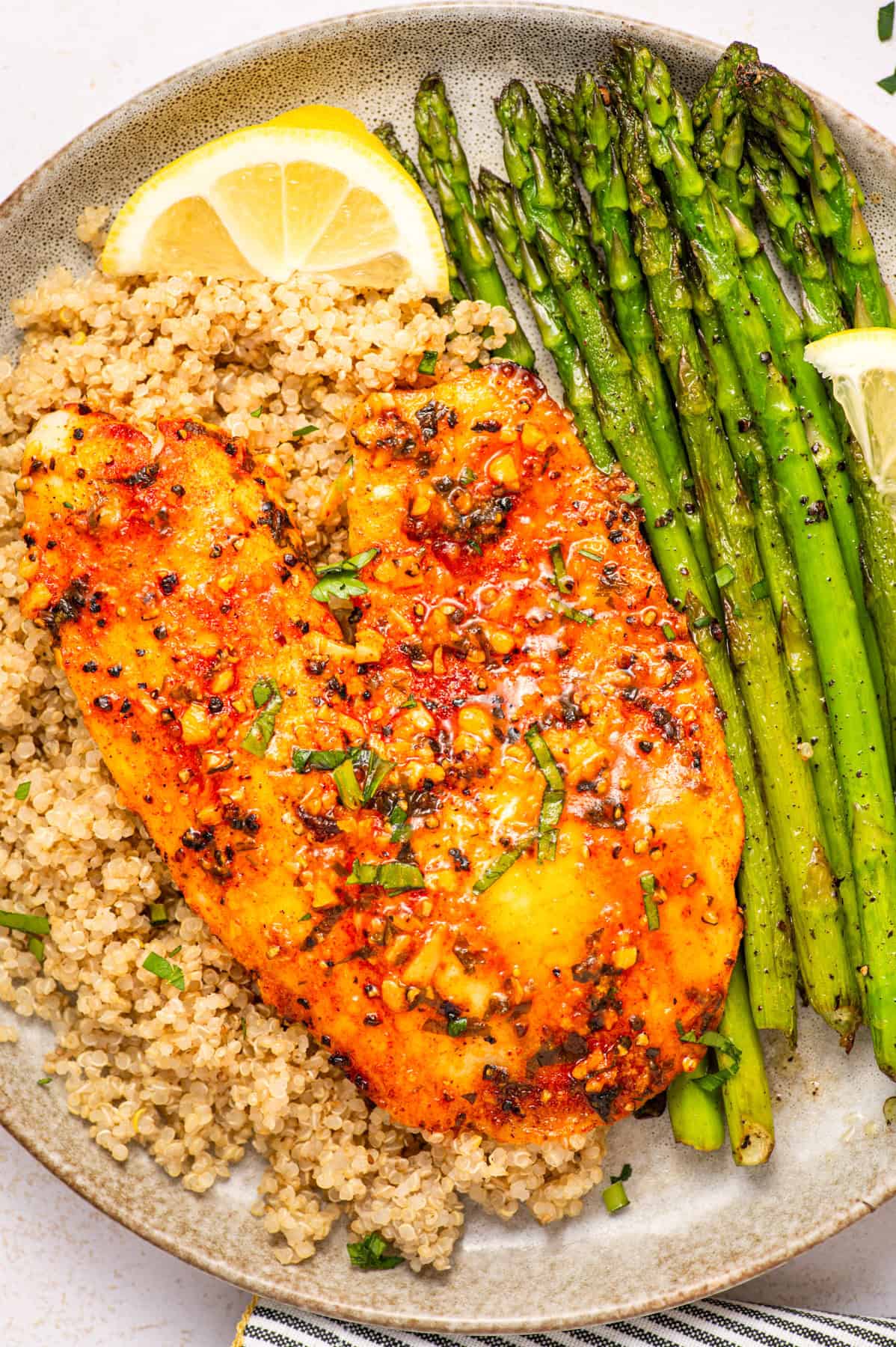 Lemon Pepper Baked Tilapia with Quinoa and Asparagus A plate with a seasoned, baked tilapia fillet on a bed of quinoa, garnished with herbs. Grilled asparagus and lemon wedges are arranged on the side.