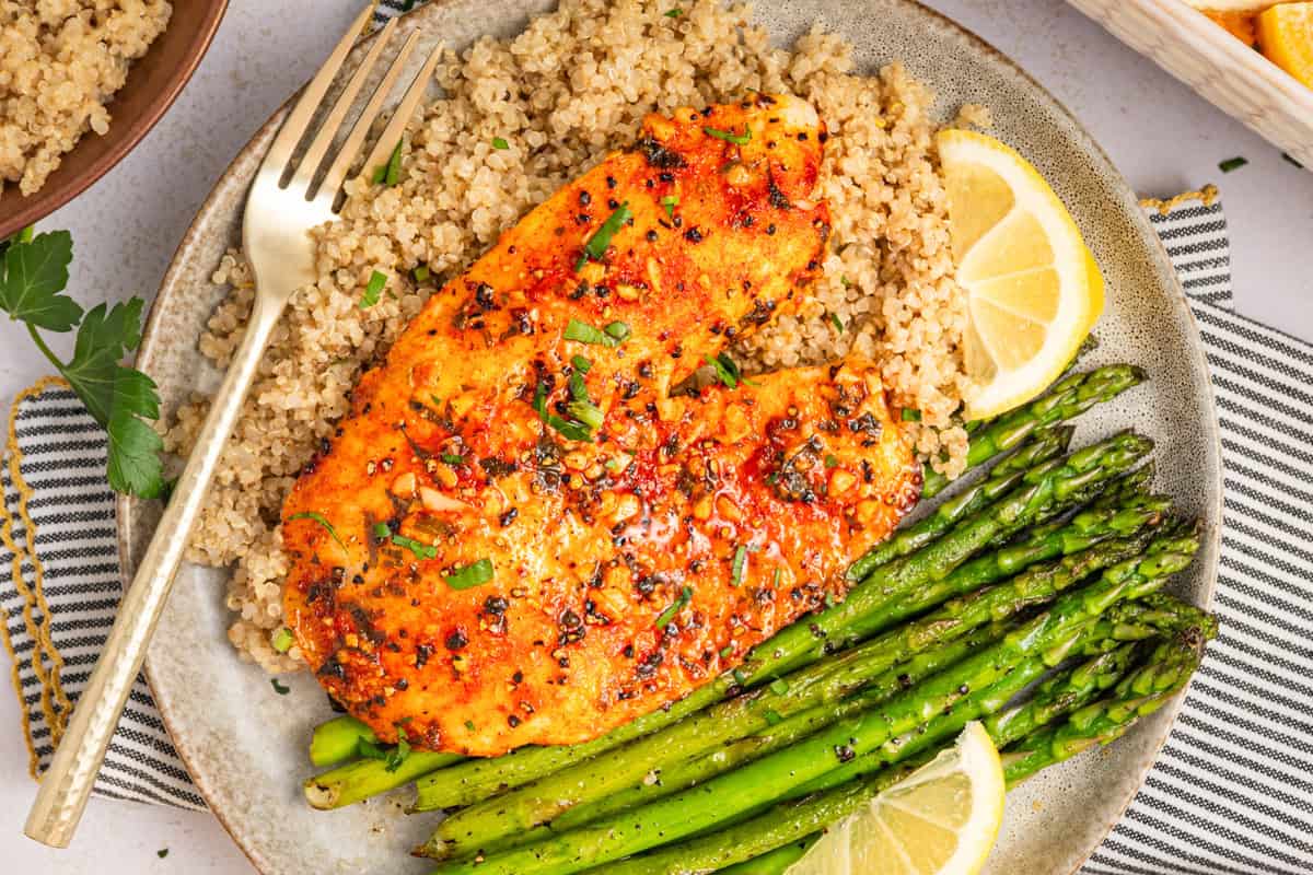 Lemon Pepper Baked Tilapia (with Lemon Butter Garlic Sauce) A plate with seasoned baked tilapia, a serving of quinoa, roasted asparagus spears, and two lemon wedges rests beside a fork. The dish is garnished with fresh herbs.