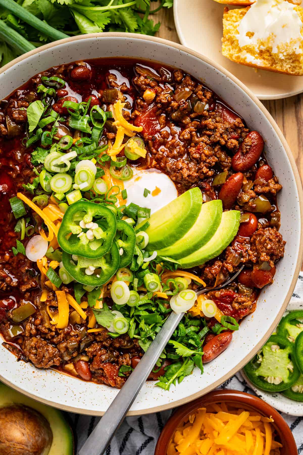 Beef Chili A bowl of chili topped with sliced avocado, jalapeños, shredded cheddar, sour cream, green onions, and cilantro. Kidney beans, ground meat, and vegetables are visible, with cornbread and butter on the side.