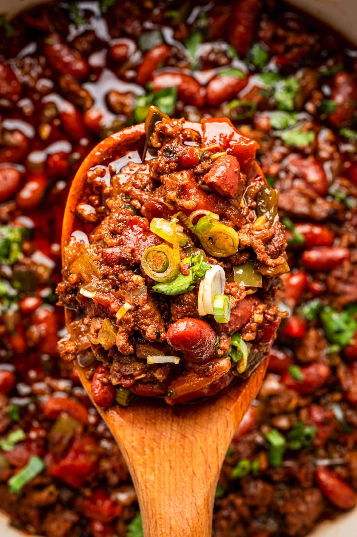 Beef Chili A close-up of a wooden spoon holding a serving of chili with ground meat, kidney beans, chopped tomatoes, green onions, and herbs, above a pot filled with more chili.
