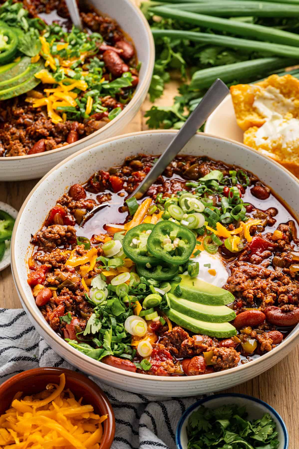 Beef Chili Two bowls of chili topped with sliced avocado, jalapeños, cheddar cheese, green onions, and cilantro. Cornbread is on the side, with fresh green onions and cilantro in the background.