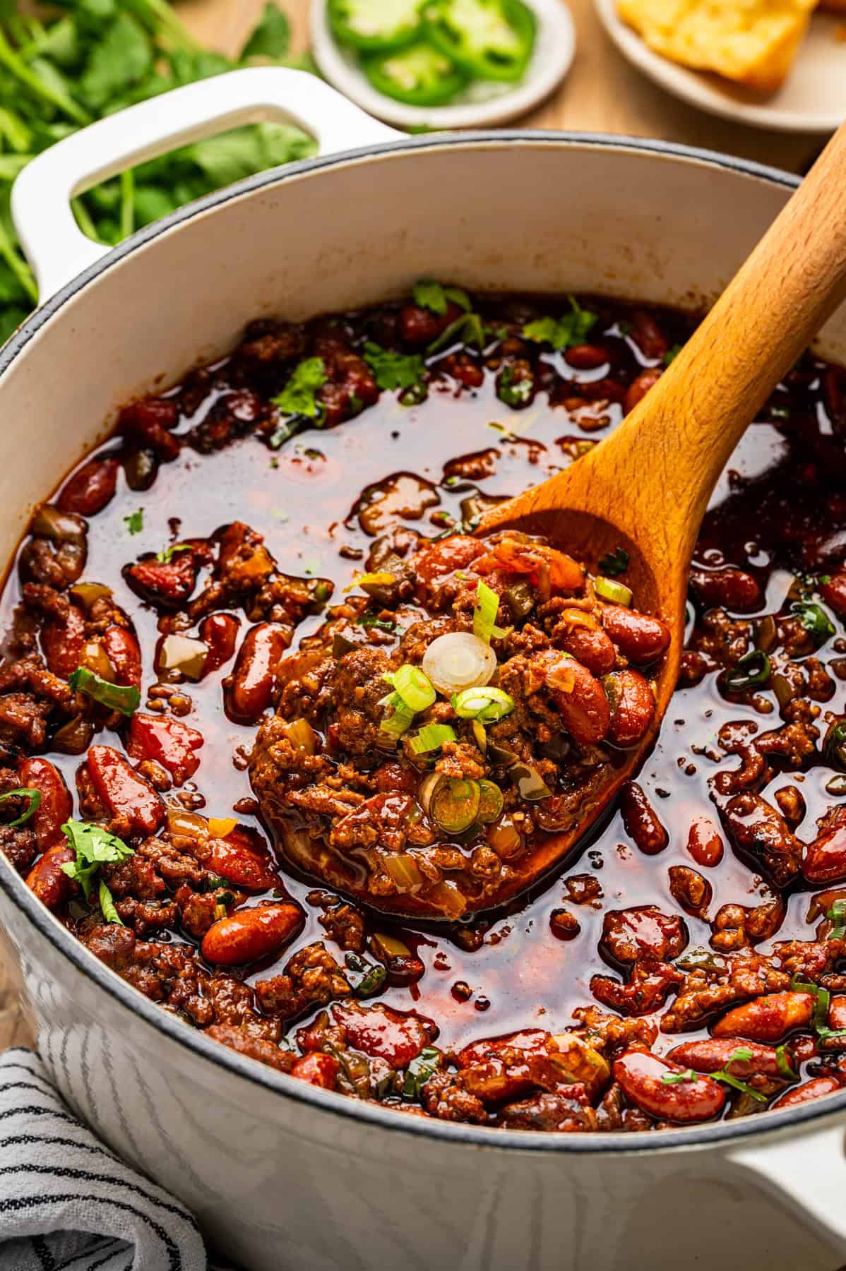 Beef Chili A pot of chili with ground meat, kidney beans, and spices, garnished with chopped green onions. A wooden spoon scoops up a hearty serving from the rich, dark, and flavorful stew.