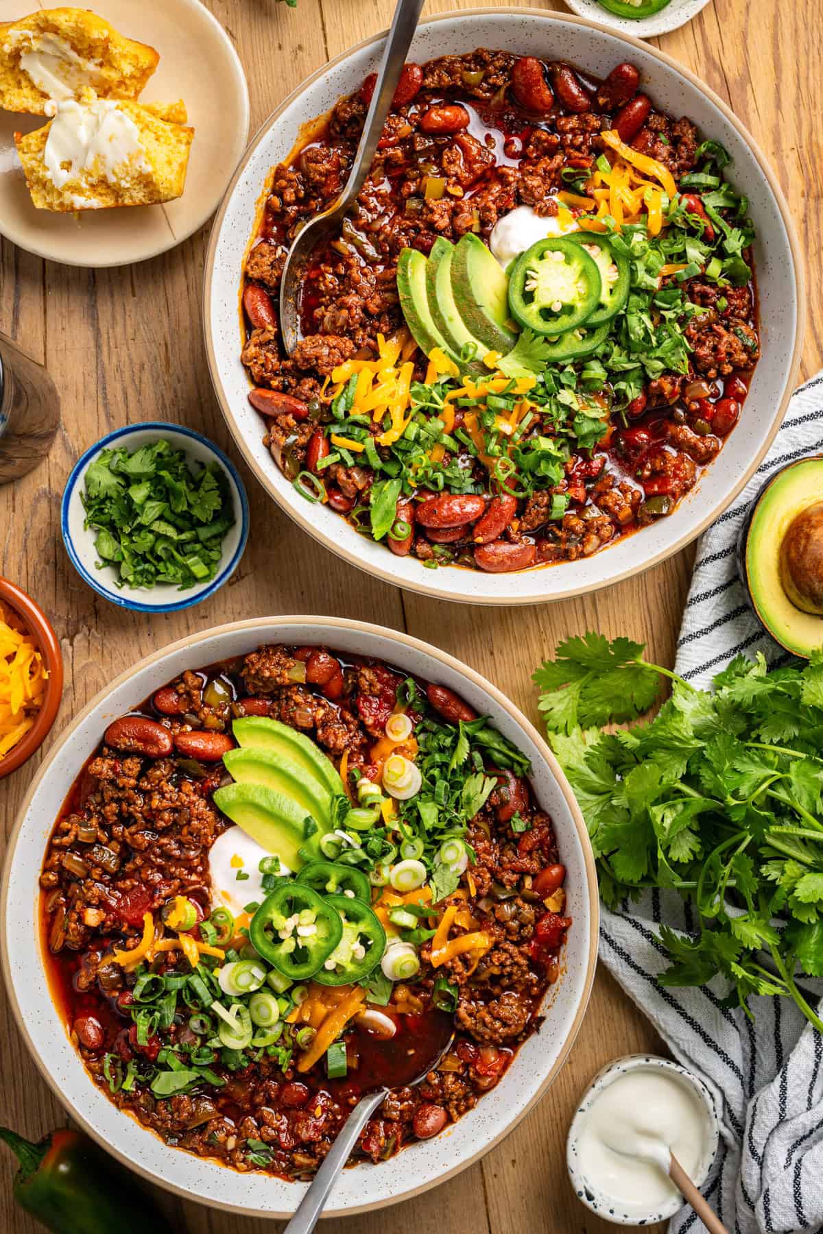 Beef Chili Two bowls of chili topped with sliced avocado, cheddar cheese, jalapeños, sour cream, and chopped green onions, surrounded by cilantro, corn muffins, shredded cheese, half an avocado, and a bowl of sour cream on a wooden table.