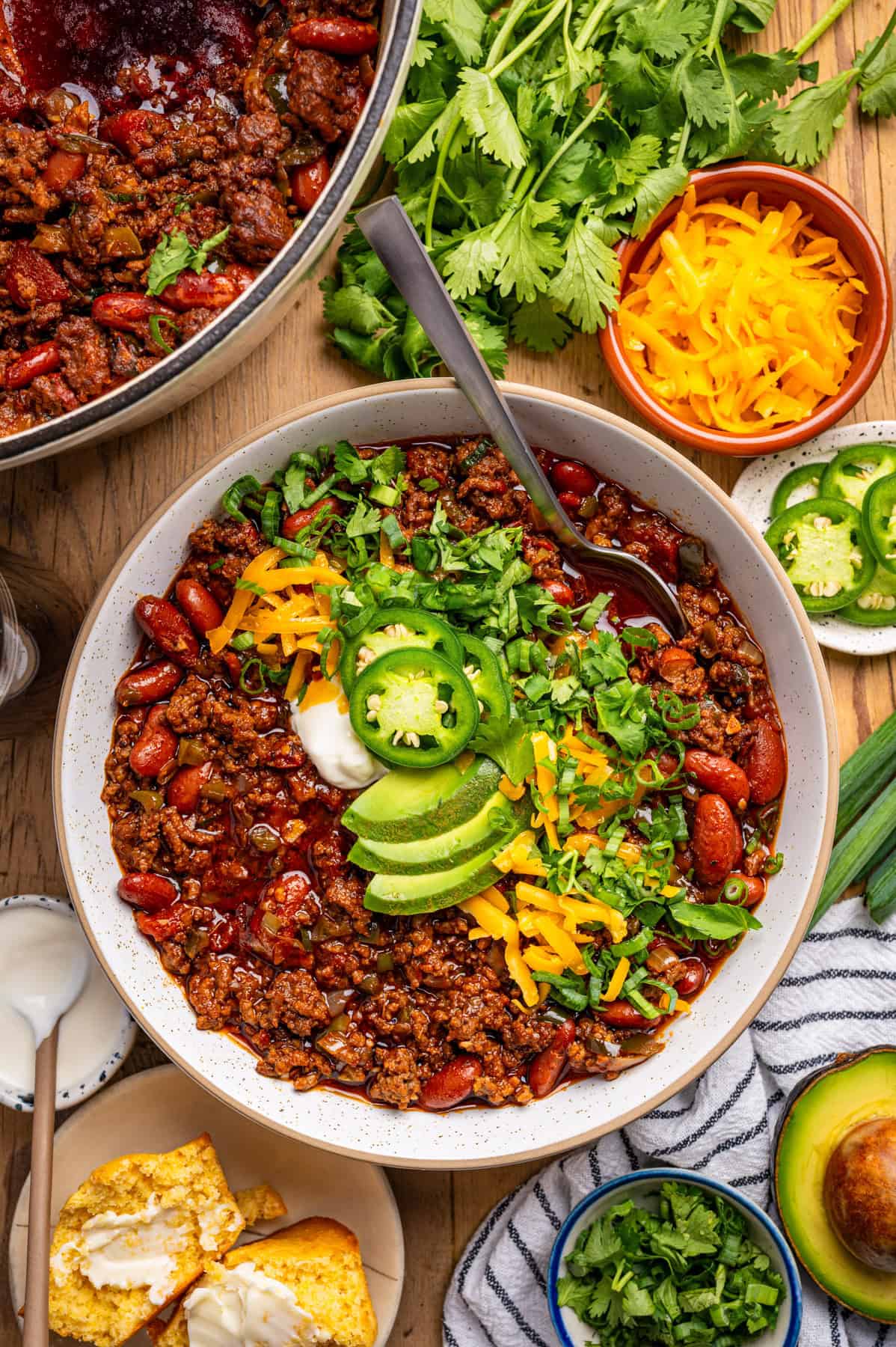 Beef Chili A bowl of beef chili topped with sliced avocado, jalapeños, shredded cheese, sour cream, and cilantro, surrounded by cornbread, green onions, more cilantro, shredded cheese, and sliced jalapeños on a wooden table.