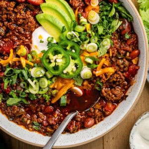 A bowl of chili topped with sliced avocado, jalapeños, shredded cheddar, green onions, cilantro, and a dollop of sour cream, with a spoon resting in the hearty, chunky chili.