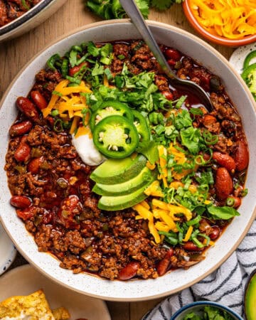 A bowl of chili with ground beef, kidney beans, and rich sauce, topped with sliced avocado, shredded cheddar, sour cream, jalapeño slices, and chopped cilantro, surrounded by ingredients and tortilla chips on a table.