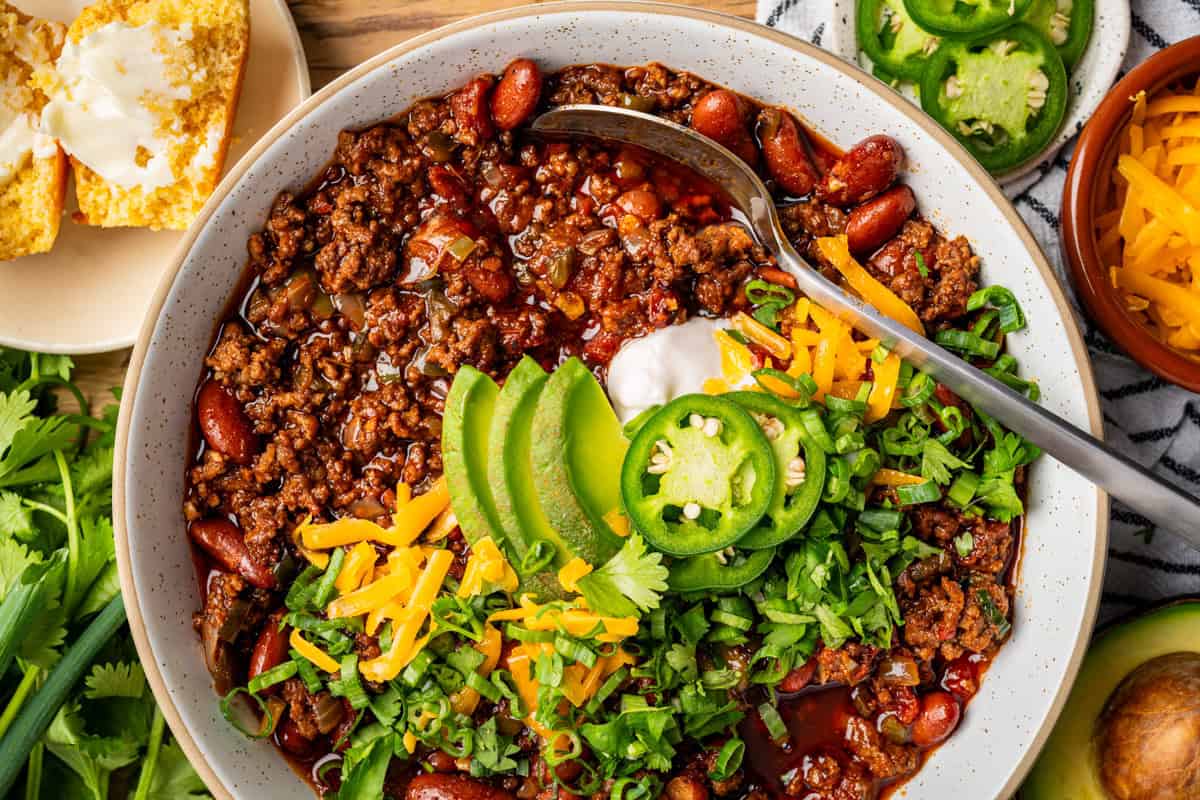 The Only Chili Recipe You'll Ever Need! A bowl of chili topped with sliced avocado, jalapeños, shredded cheese, sour cream, and chopped cilantro. Kidney beans and ground beef are visible. Cornbread with butter is on a plate nearby, with herbs and a sliced jalapeño on the side.