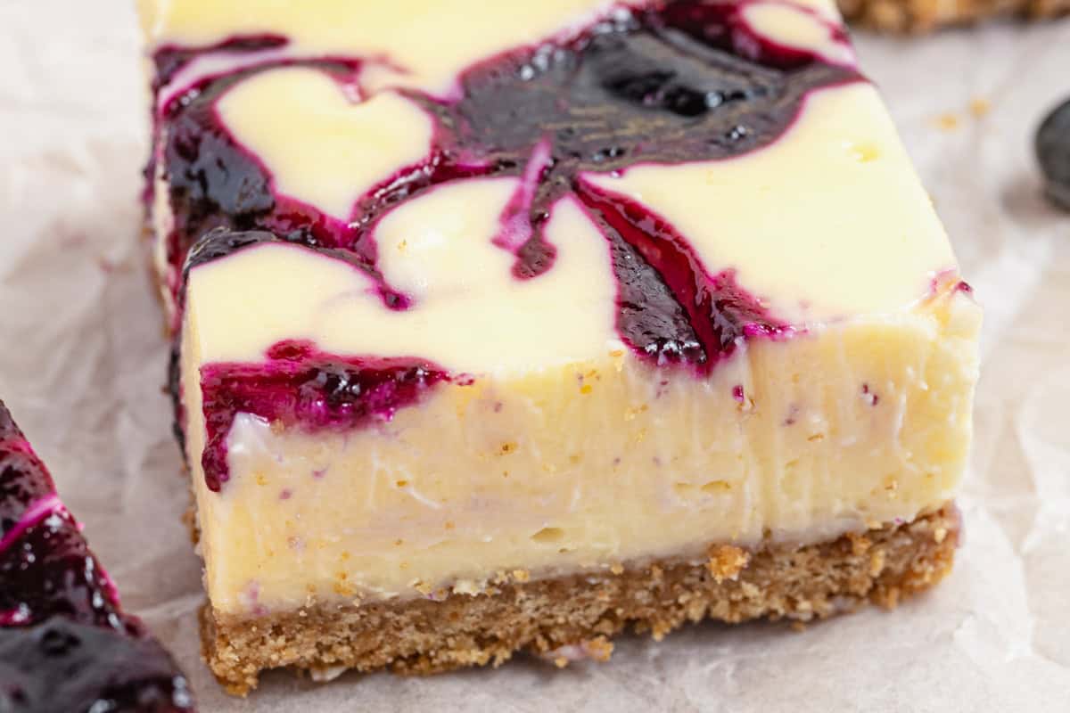 A close-up of a cheesecake bar with a crumbly brown crust, creamy filling, and swirls of dark purple blueberry sauce on top, sitting on a piece of parchment paper.