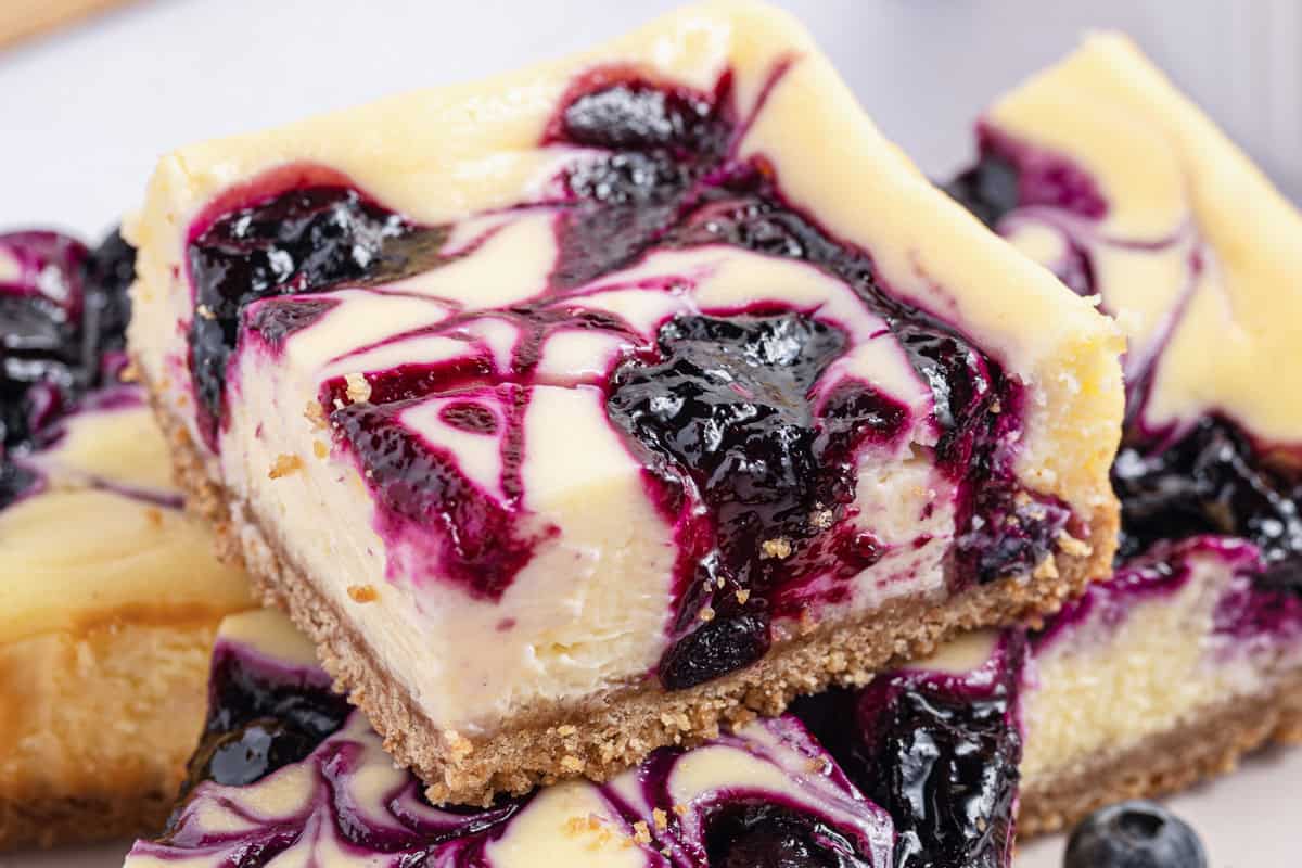 A close-up of a cheesecake bar with a graham cracker crust, creamy filling, and a vibrant blueberry swirl on top.