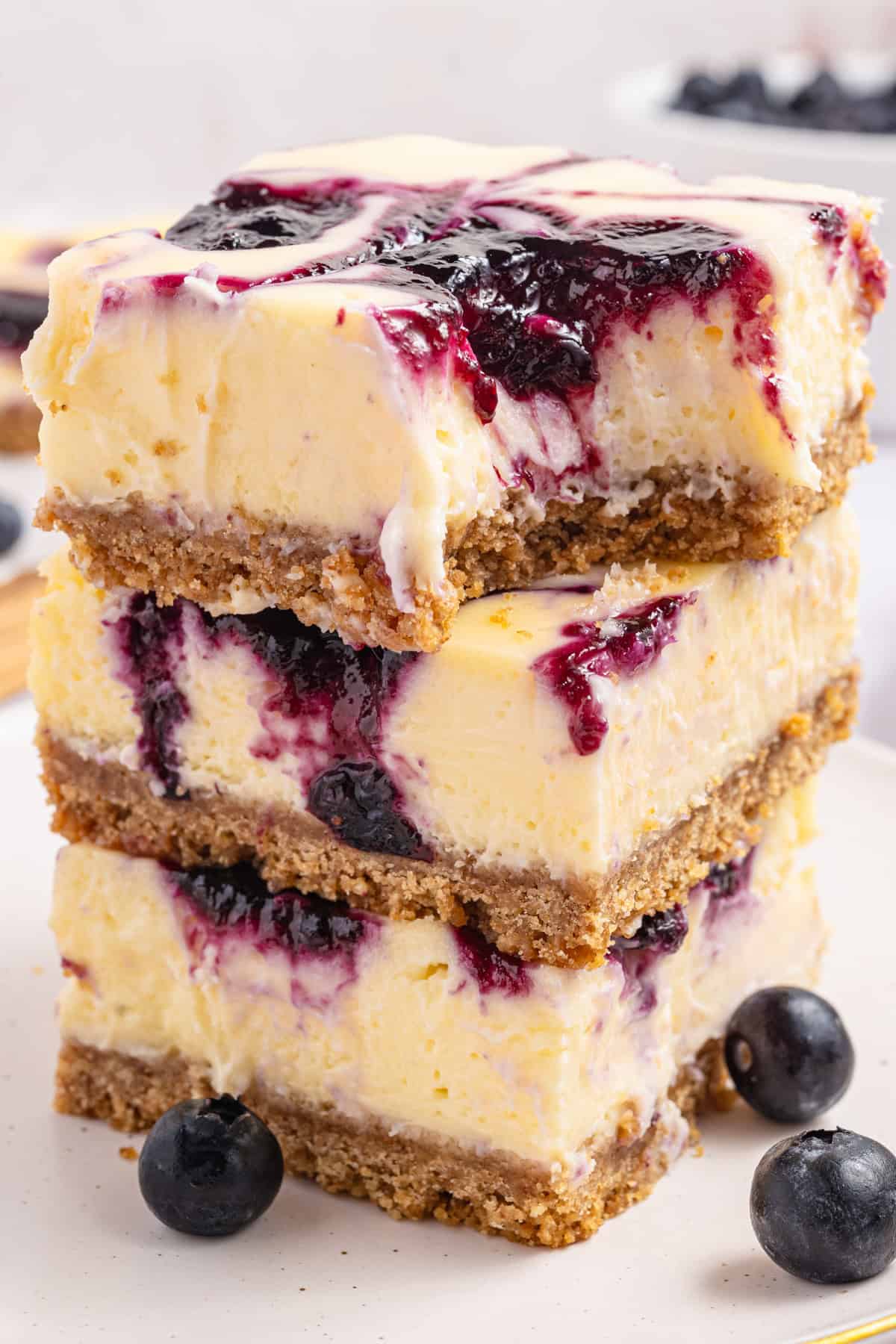 Three thick blueberry cheesecake bars are stacked on a plate. Each bar has a graham cracker crust, creamy cheesecake layer, and blueberry swirl topping, with fresh blueberries scattered around.