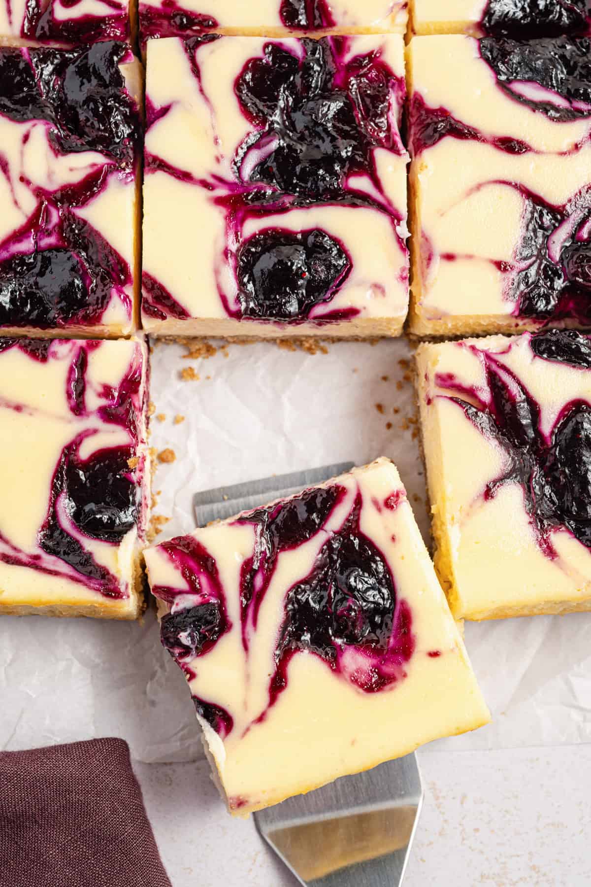 Close-up of creamy cheesecake bars with a dark purple blueberry swirl, cut into squares. One piece is being lifted with a spatula, revealing a crumbly crust underneath. The bars rest on white parchment paper.