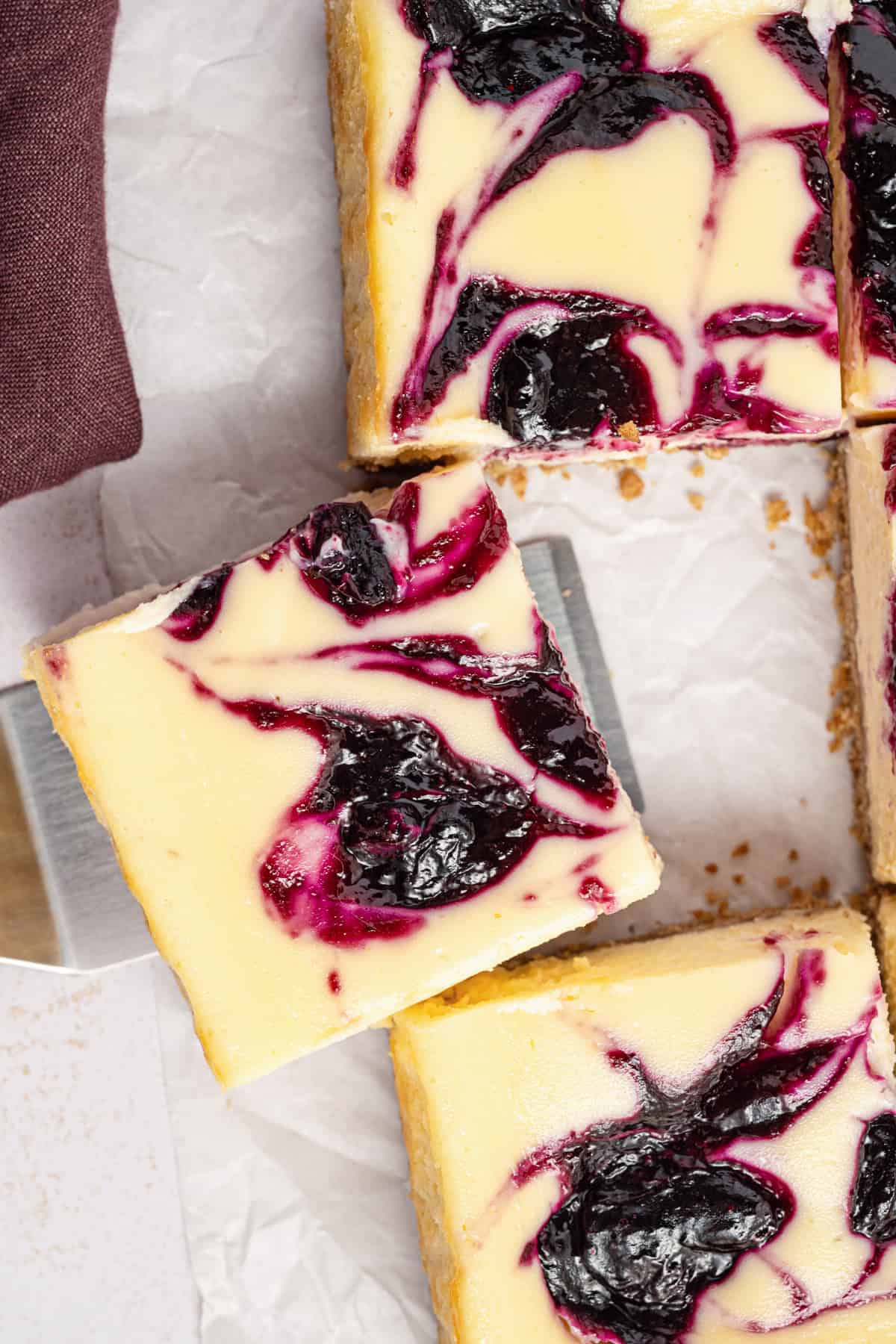A close-up of cheesecake bars topped with swirls of dark berry sauce, arranged on parchment paper. One bar is being lifted with a spatula. The bars have a creamy texture and vibrant berry patterns.