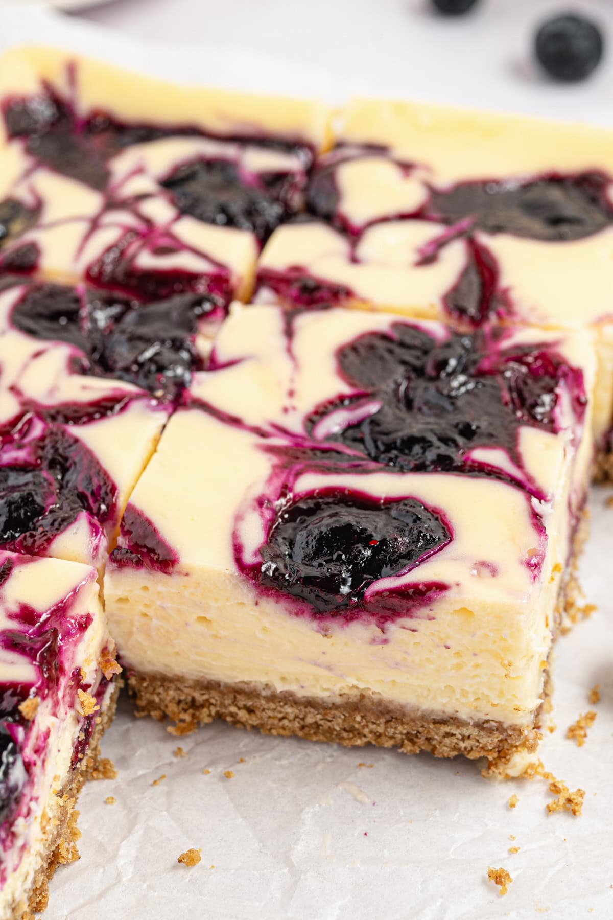 Cheesecake bars with a graham cracker crust, topped with a blueberry swirl. The bars are cut into squares, revealing creamy layers and vibrant purple-blue berry filling on top.