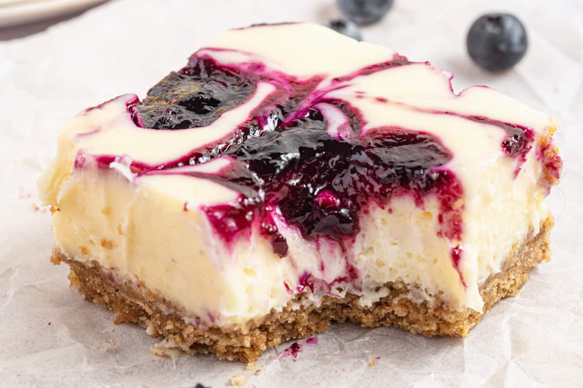 A close-up of a cheesecake bar with a crumbly crust and creamy filling, topped with a swirl of blueberry sauce. Two blueberries are blurred in the background.