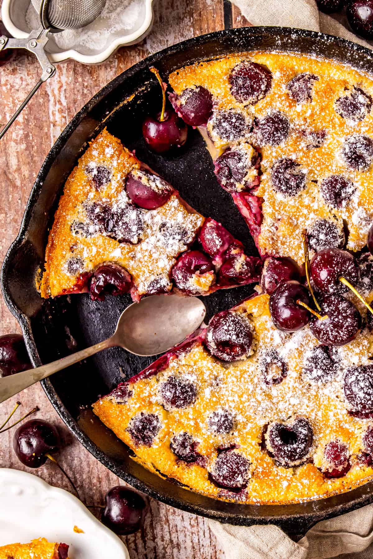 A cherry clafoutis baked in a cast iron skillet, dusted with powdered sugar. Two slices are cut, one slightly separated with a spoon beside it. Fresh cherries are scattered on top and around the skillet on a wooden table.