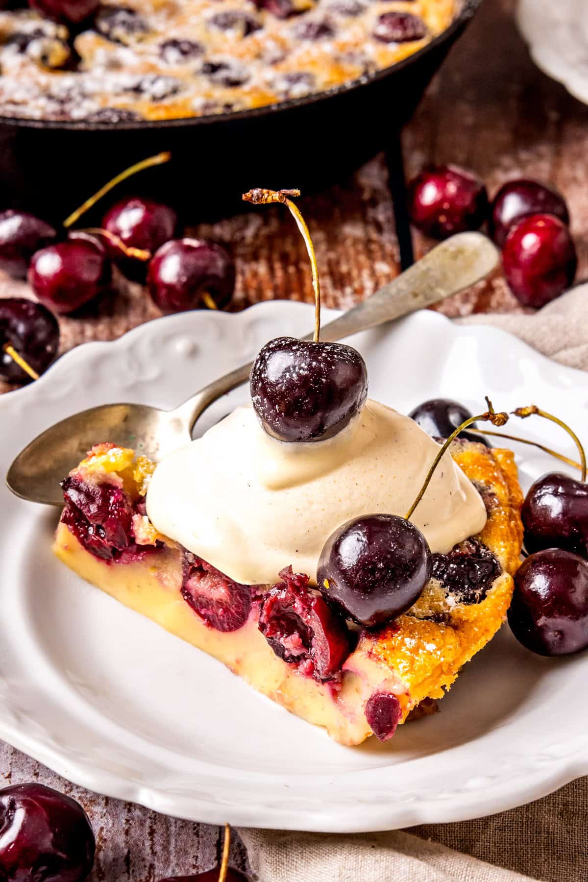 A slice of cherry clafoutis topped with a scoop of vanilla ice cream and fresh cherries sits on a white plate, surrounded by more cherries and a spoon, with additional clafoutis in the background.