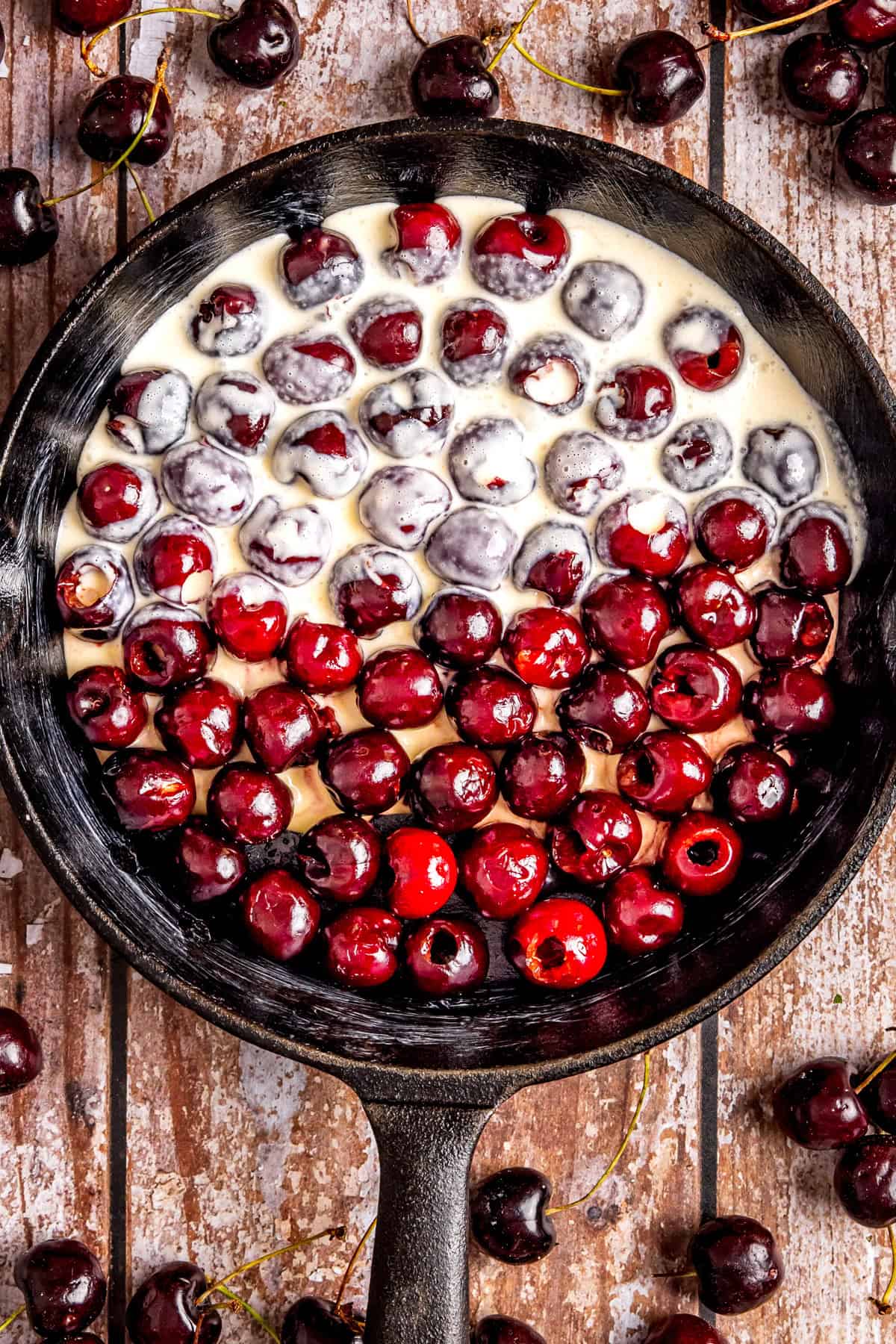 A cast-iron skillet filled with fresh cherries, some covered in a clafoutis batter, sits on a rustic wooden surface with more cherries scattered around.