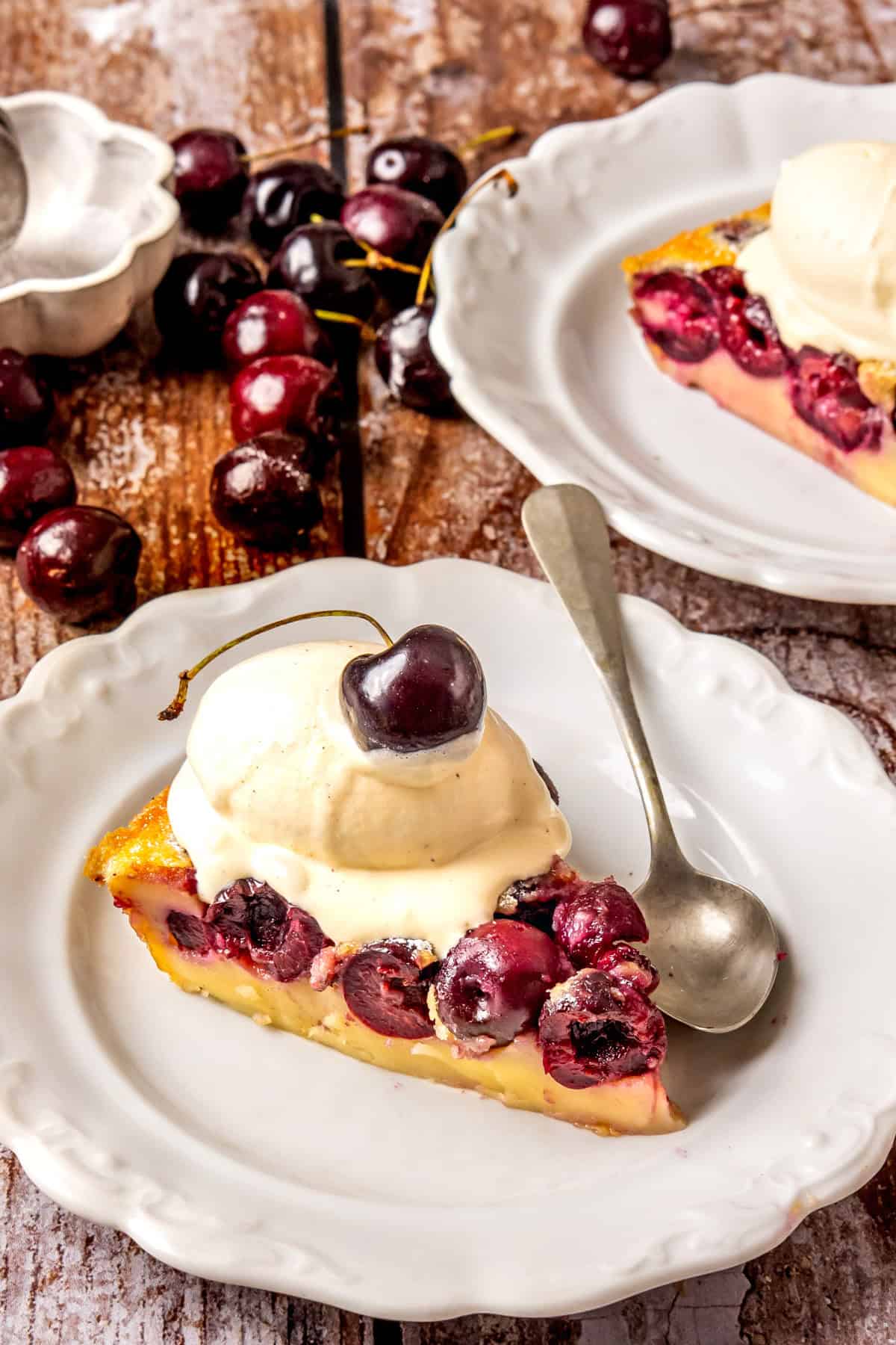 A slice of cherry clafoutis topped with a scoop of vanilla ice cream and a cherry on a white plate, with a spoon beside it. Fresh cherries and another plate with dessert are in the background on a rustic wooden table.