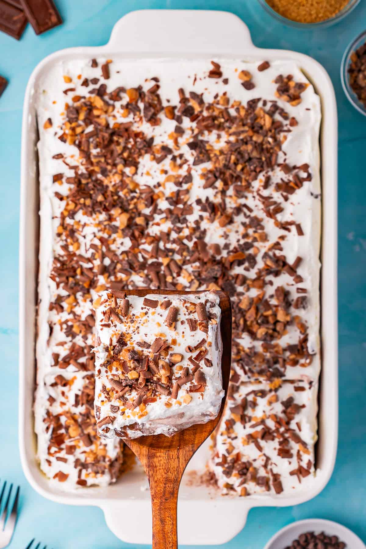 A wooden spatula lifts a square piece of chocolate delight-a whipped cream dessert topped with chocolate and toffee bits-from a tray filled with the same treat. Bowls with toppings and utensils are visible nearby on a blue surface.