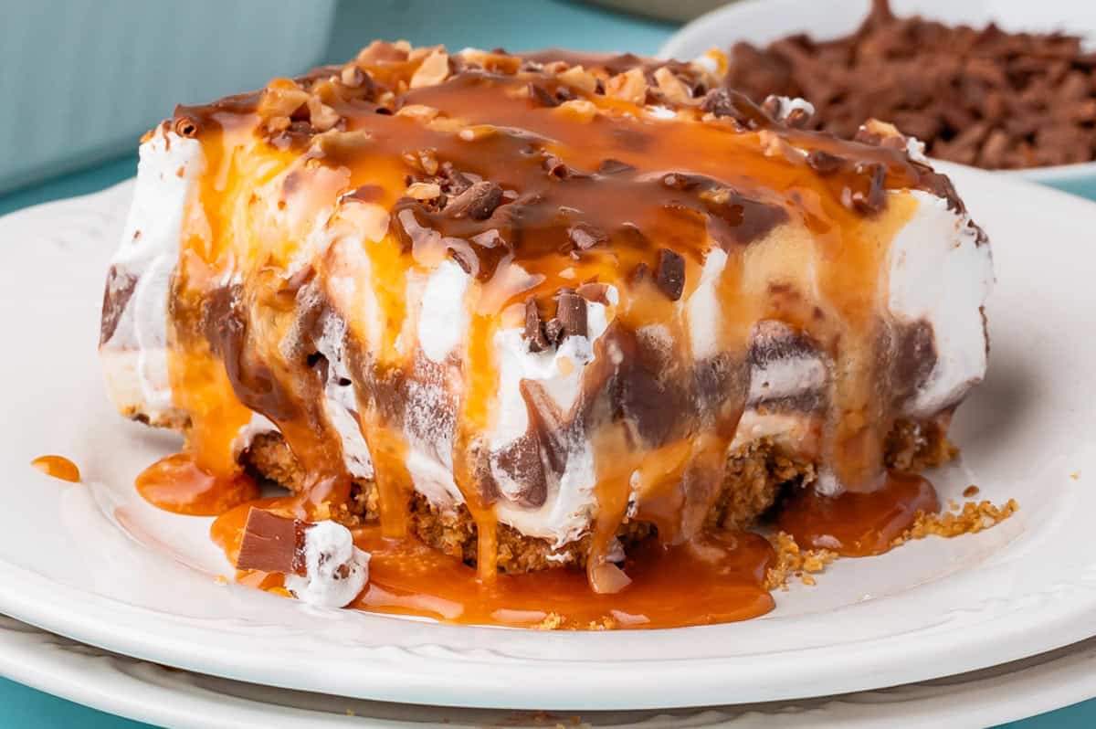A close-up of chocolate delight: a layered dessert with a graham cracker crust, chocolate and vanilla pudding, whipped topping, chocolate shavings, and caramel sauce drizzled on top, served on a white plate.