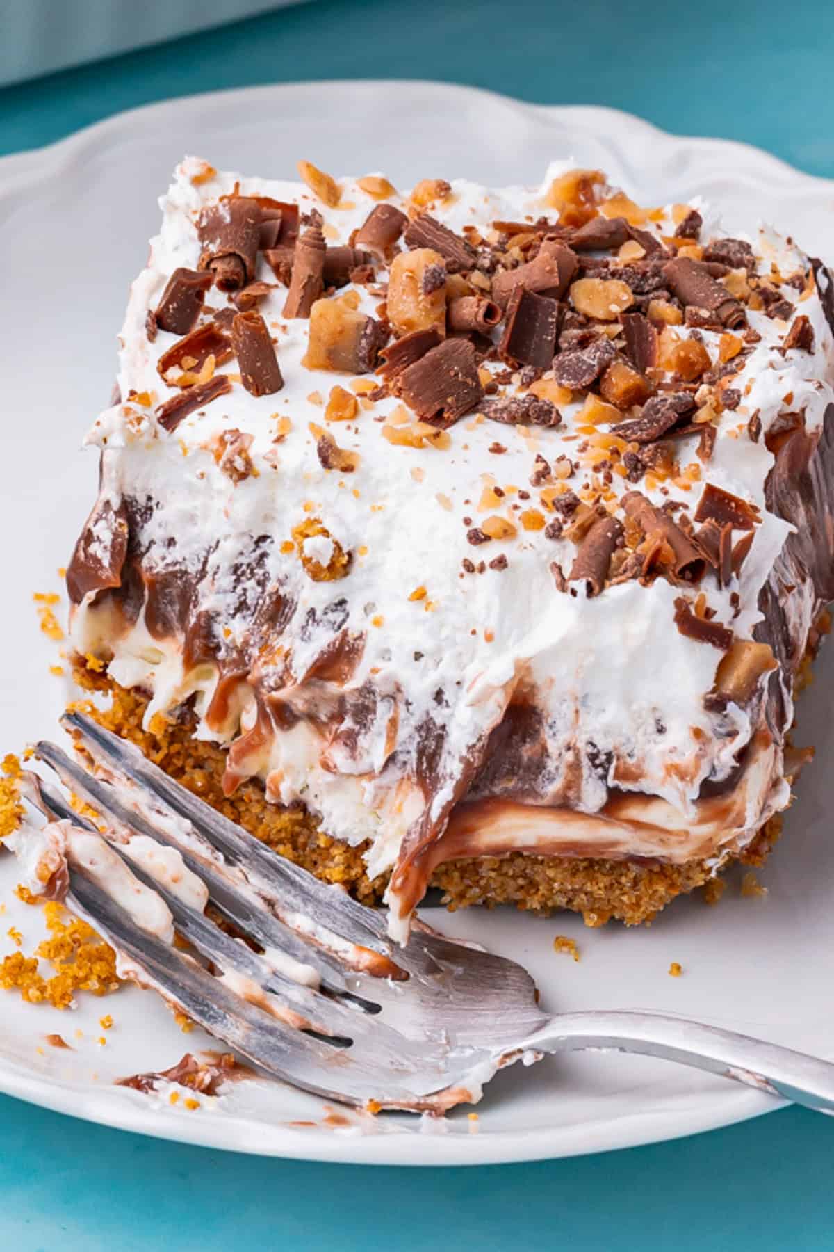A close-up of a chocolate delight dessert slice with a graham cracker crust, creamy layers, chocolate pudding, whipped topping, and chopped candy pieces. A fork rests on the plate with crumbs and cream.