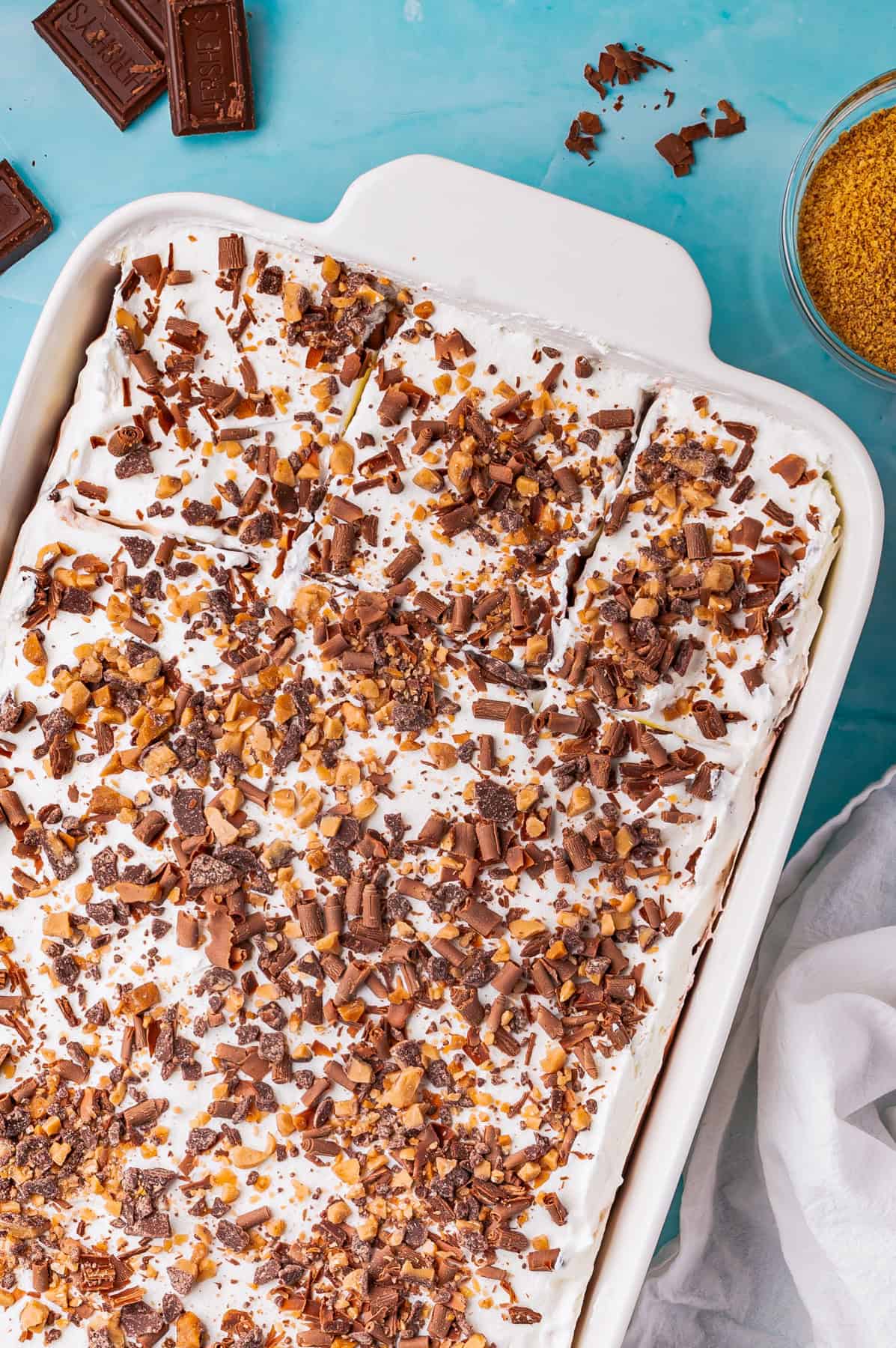 A rectangular dish filled with chocolate delight, topped with whipped cream and sprinkled with chopped chocolate and toffee bits, surrounded by a few chocolate squares and a bowl of brown sugar on a blue surface.