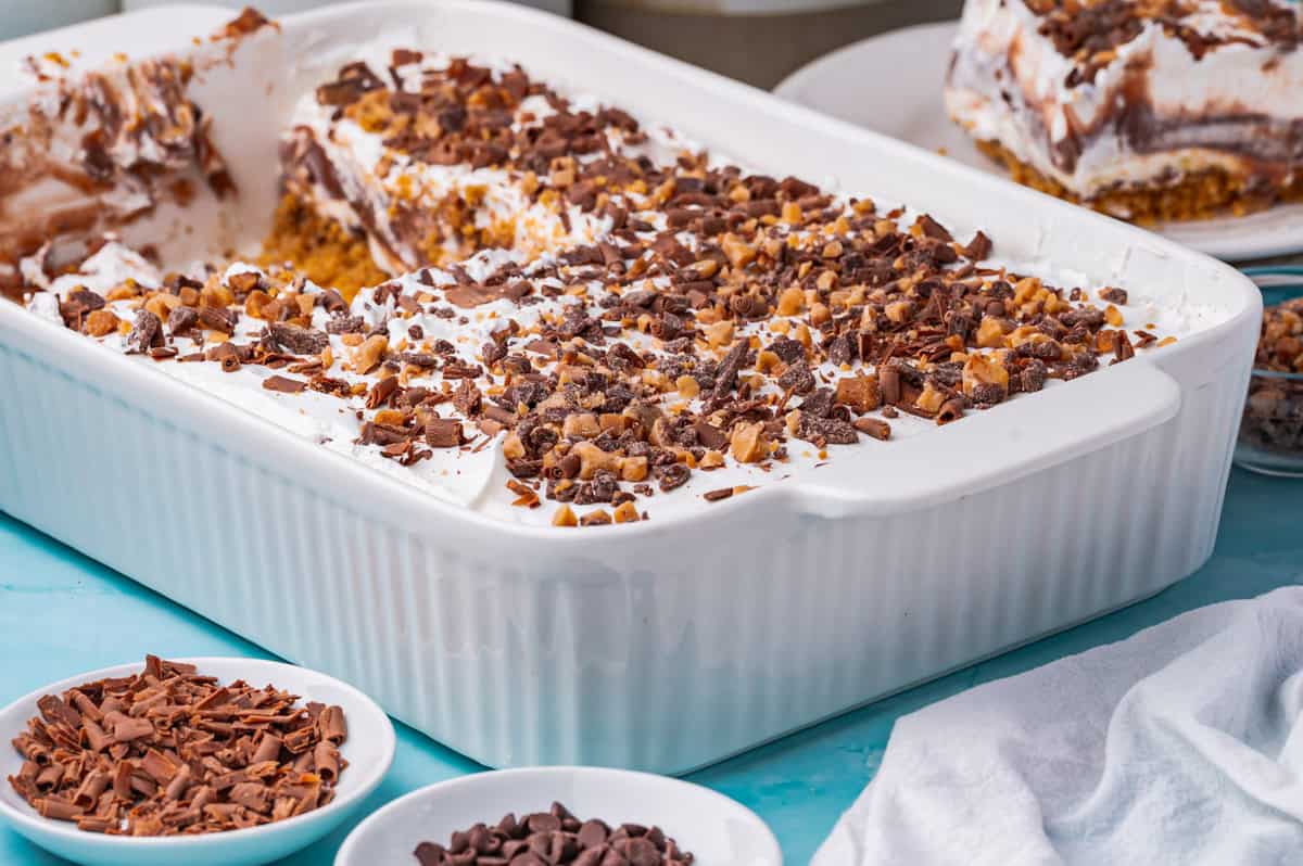 A large white baking dish filled with chocolate delight, this layered dessert is topped with whipped cream and chocolate shavings, with slices served on plates and bowls of chocolate chips nearby on a blue surface.