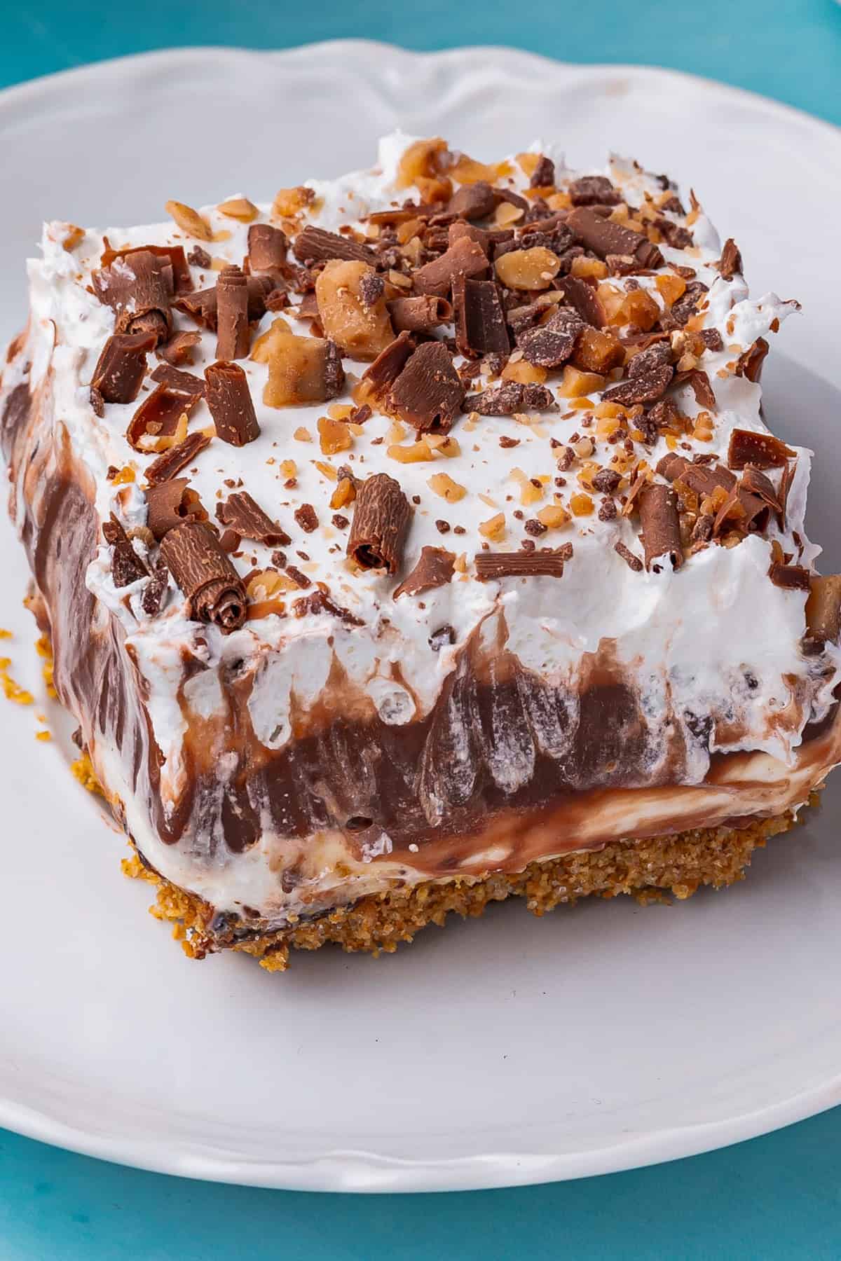 A close-up of a chocolate delight dessert slice with layers of graham cracker crust, creamy filling, chocolate pudding, whipped topping, and a sprinkle of chocolate shavings and toffee bits on a white plate.