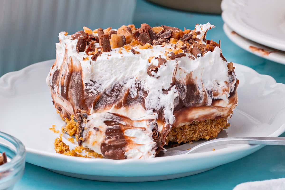 A slice of chocolate delight with a graham cracker crust, creamy chocolate and vanilla layers, topped with whipped cream and chocolate shavings, served on a white plate with a fork.