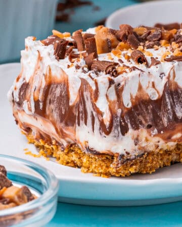 A close-up of a creamy layered chocolate delight with a crumbly crust, rich chocolate and whipped cream layers, topped with chocolate shavings and toffee bits, served on a white plate.