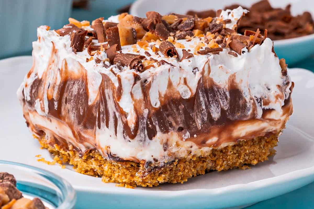 A close-up of a chocolate delight with a crumbly golden crust, layers of chocolate and creamy white filling, topped with whipped cream and chocolate shavings, served on a white plate.