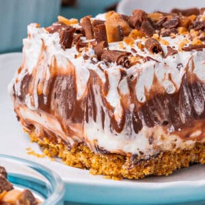 A close-up of a creamy chocolate delight with swirled chocolate and whipped cream layers on a crumbly cookie crust, topped with chocolate shavings and toffee bits, served on a white plate.