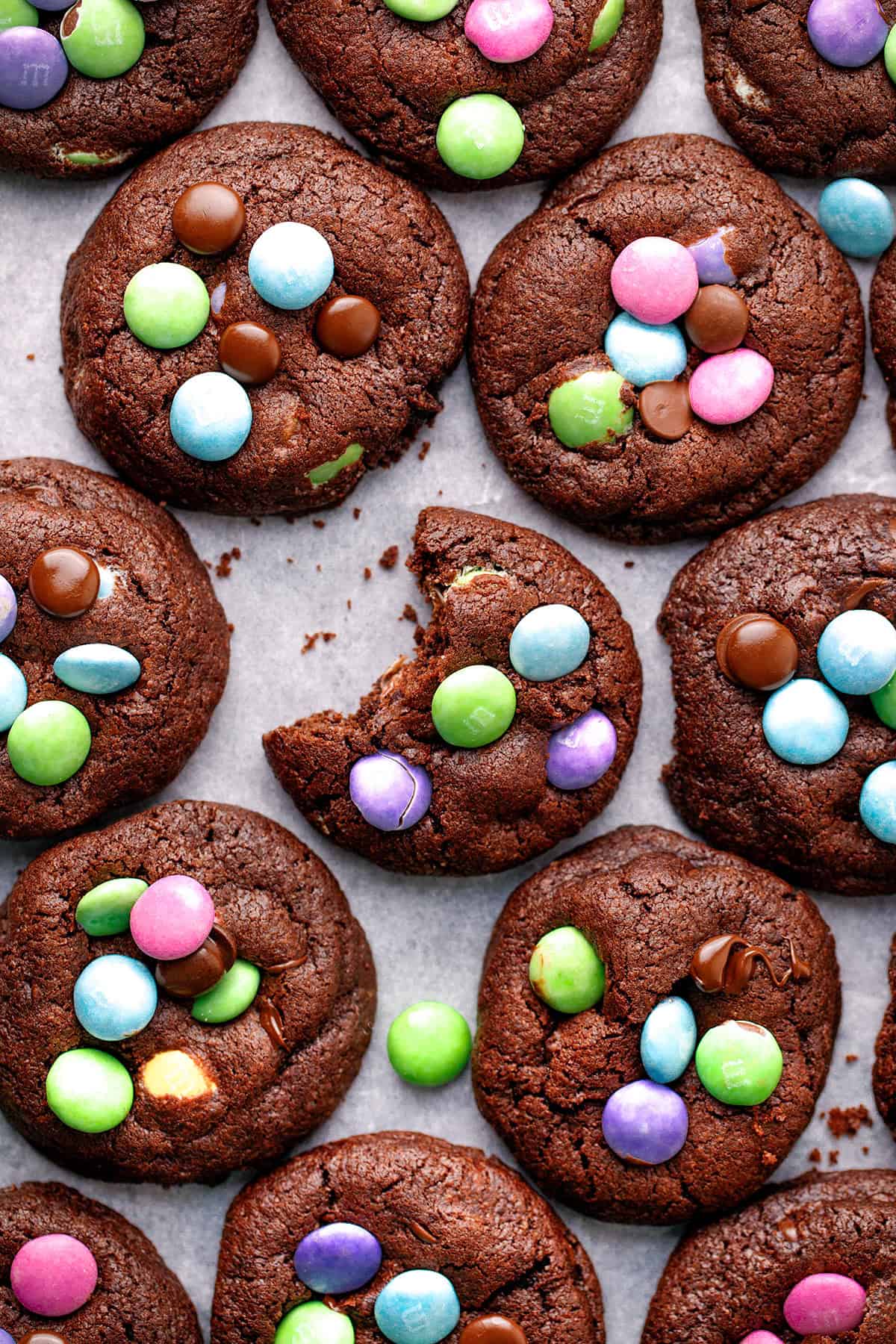 Chocolate cookies topped with colorful candy-coated chocolates are arranged in rows on parchment paper; one cookie has a bite taken out of it, revealing a soft interior.