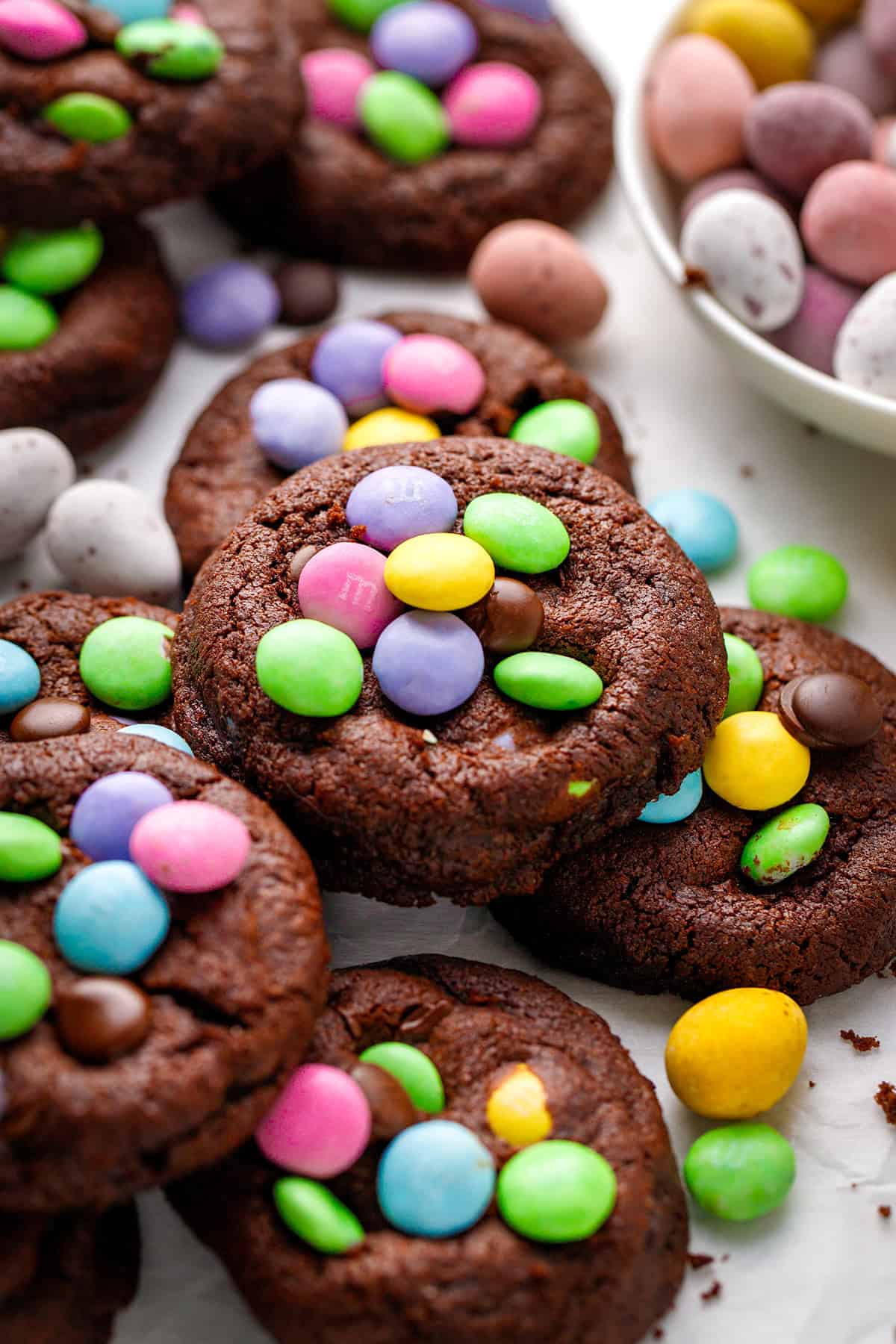 A close-up of chocolate cookies topped with colorful candy-coated chocolates, with a bowl of similar candies in the background. 
