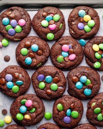 A tray of chocolate cookies topped with colorful candy-coated chocolates and chocolate chips, arranged neatly on parchment paper.