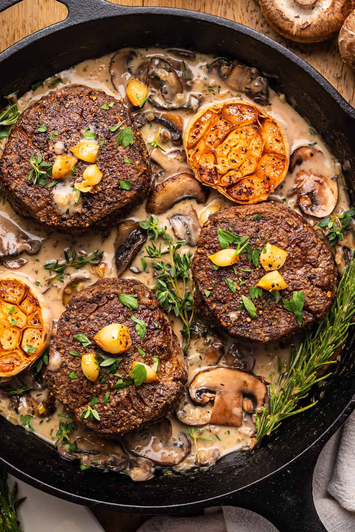 Three seared filet mignon steaks in a creamy mushroom sauce, topped with herbs and garlic, cooked in a cast iron skillet alongside fresh thyme, rosemary, and roasted tomatoes.
