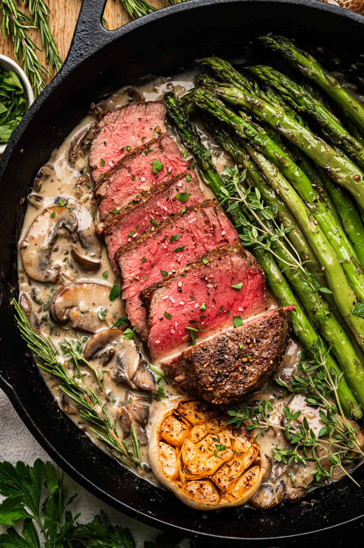 A skillet with sliced medium-rare filet mignon steak, asparagus spears, creamy mushroom sauce, fresh herbs, and a halved roasted garlic bulb, all garnished with chopped parsley.