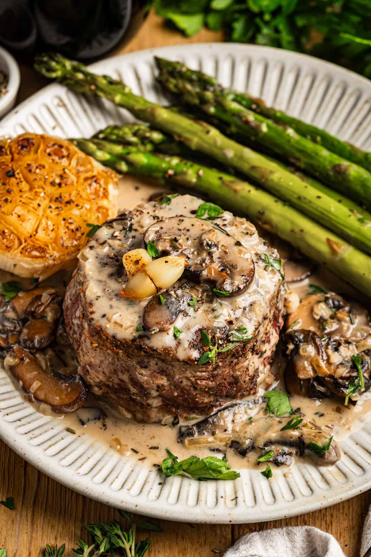 Filet mignon topped with creamy mushroom sauce, garnished with herbs and roasted garlic, served with grilled asparagus and half a roasted garlic bulb on a white plate.