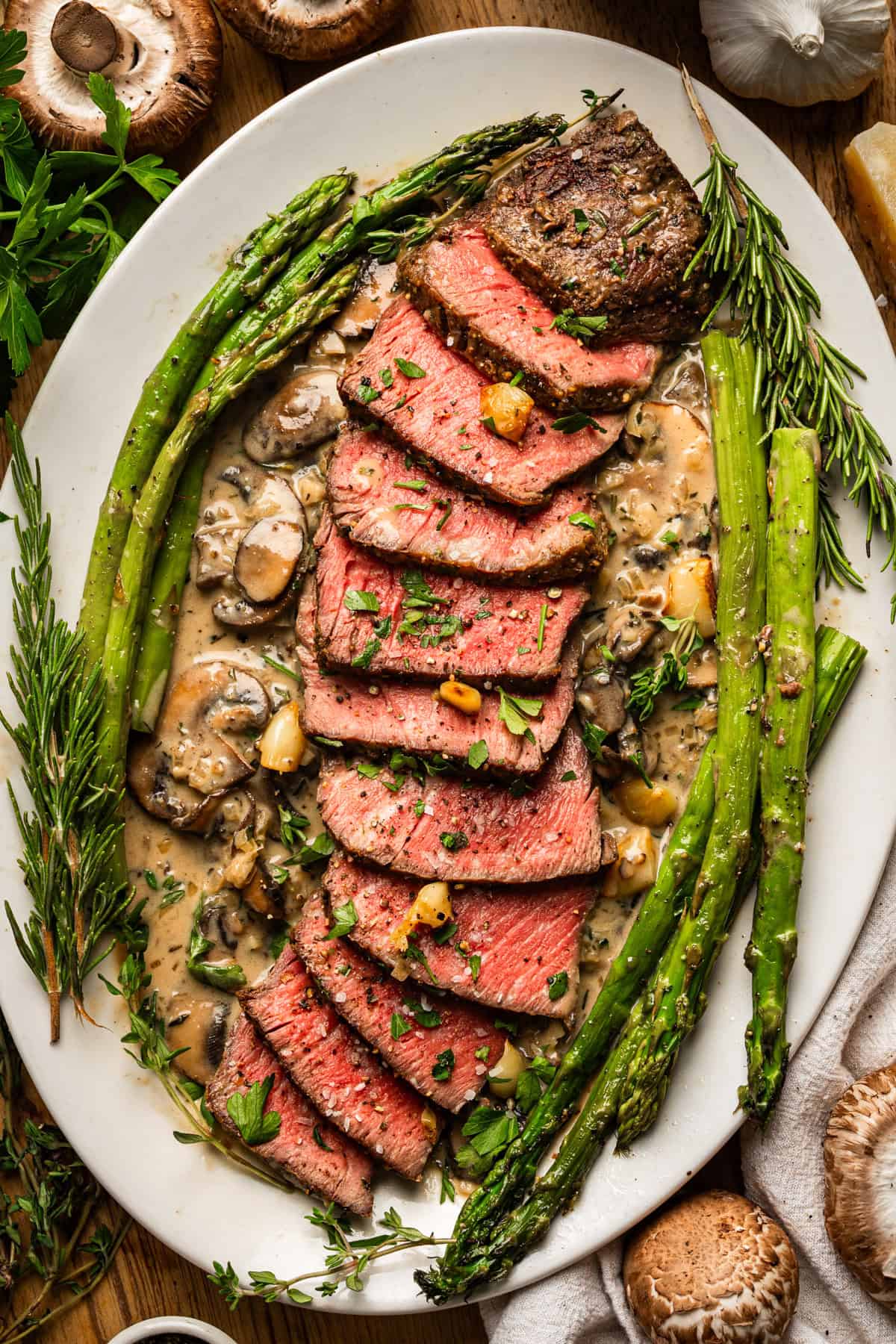 A platter of medium-rare sliced filet mignon steak topped with herbs, served on a creamy mushroom sauce with whole asparagus spears, garnished with fresh rosemary and parsley.