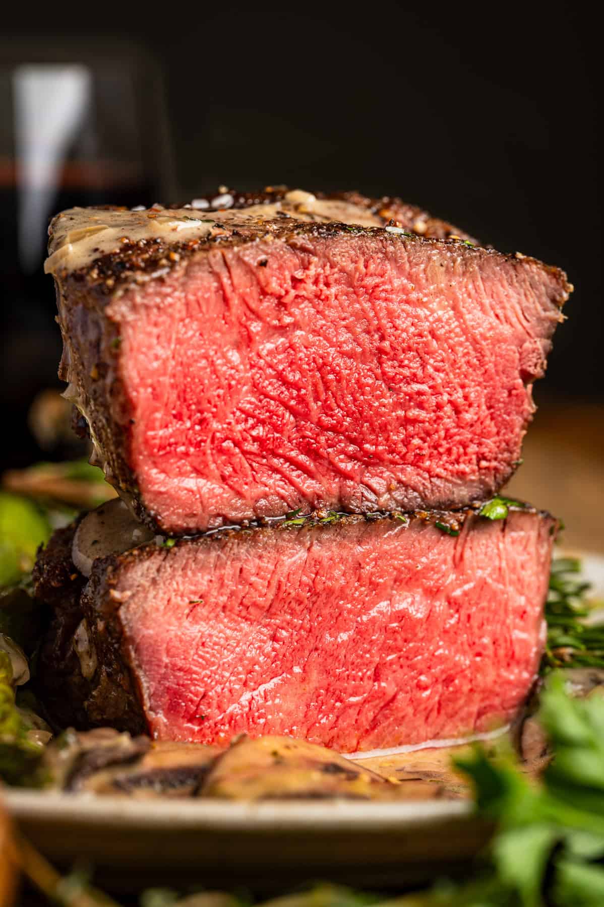 A close-up of two thick, medium-rare filet mignon steak cuts stacked on a plate, showing a juicy red center with a seared crust. Fresh herbs and mushrooms surround the steaks.
