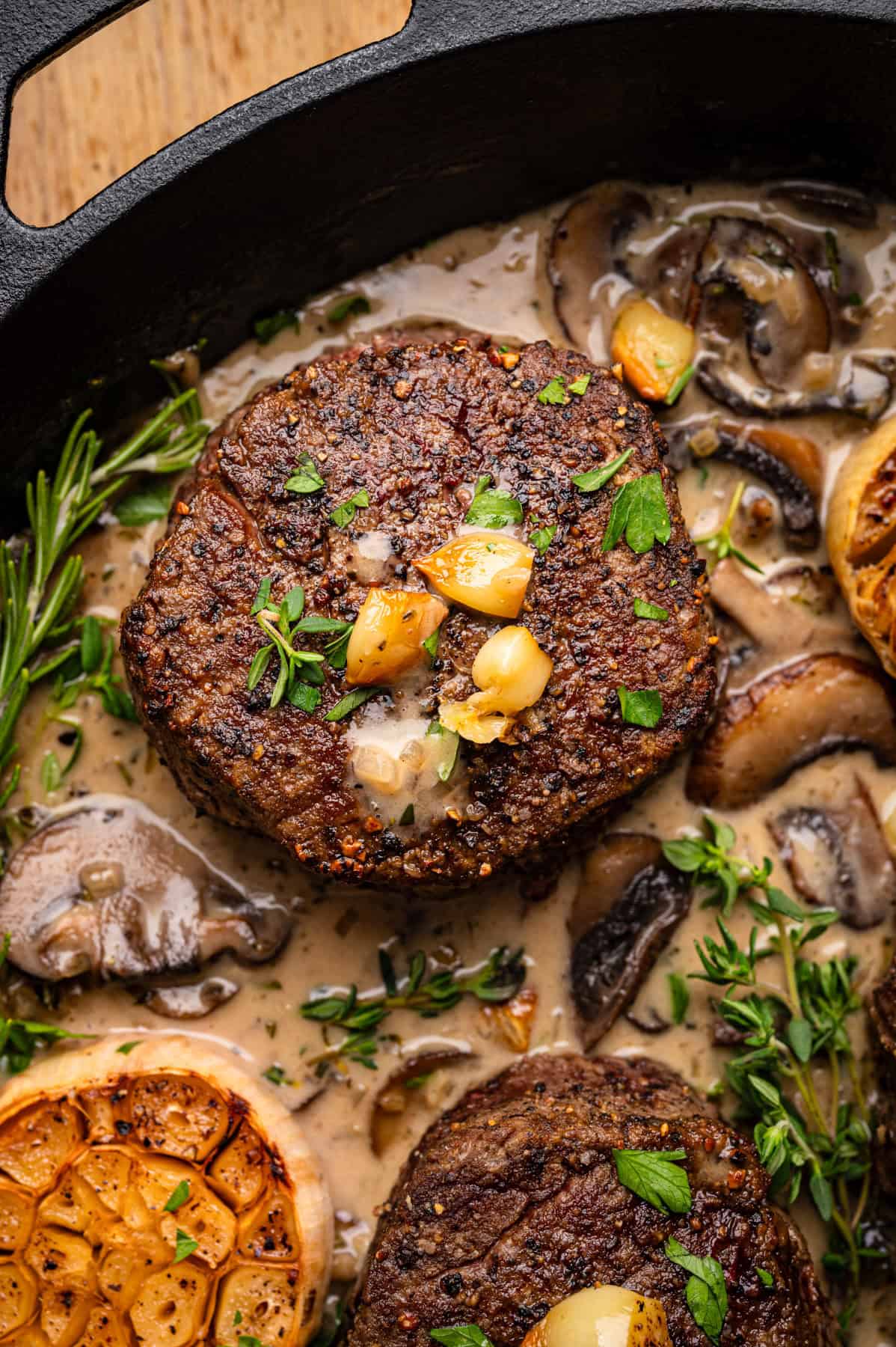 A close-up of seared filet mignon steak medallions in a creamy mushroom sauce, garnished with roasted garlic cloves, fresh herbs, and surrounded by halved roasted garlic bulbs in a cast iron skillet.