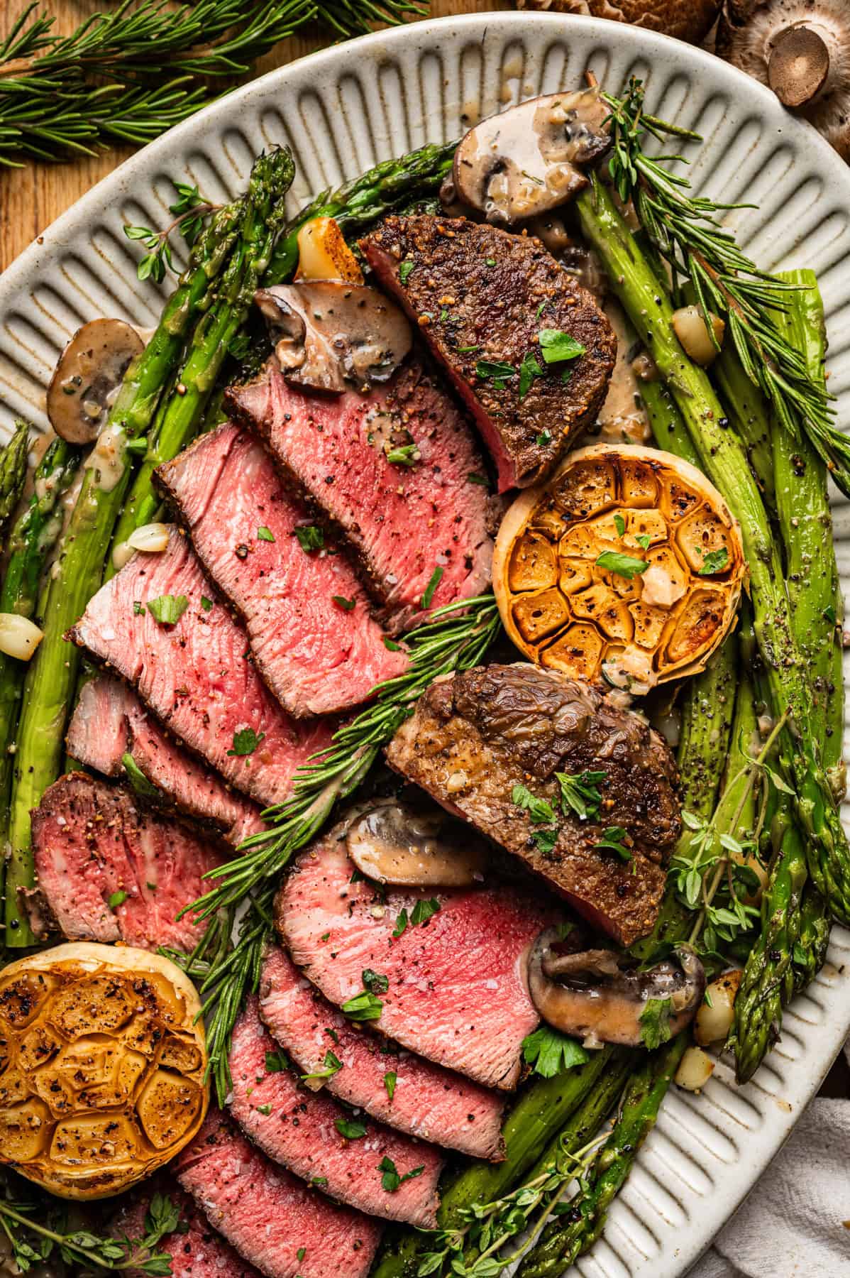 Sliced medium-rare filet mignon steak topped with herbs, surrounded by roasted asparagus, whole garlic halves, mushrooms, and fresh rosemary on a white oval plate.