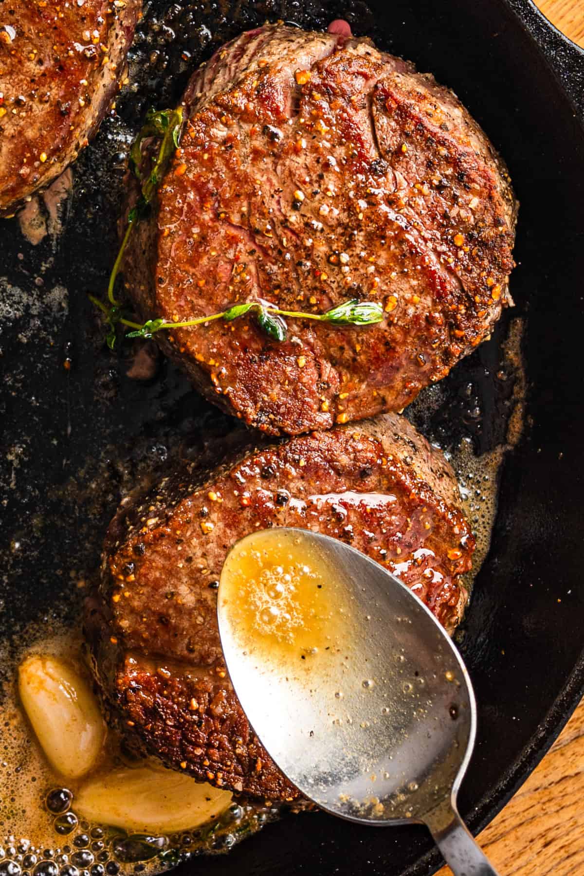Two seasoned filet mignon steaks are searing in a cast iron skillet with sprigs of thyme, garlic cloves, and melted butter, while a spoon drizzles butter over the steaks.